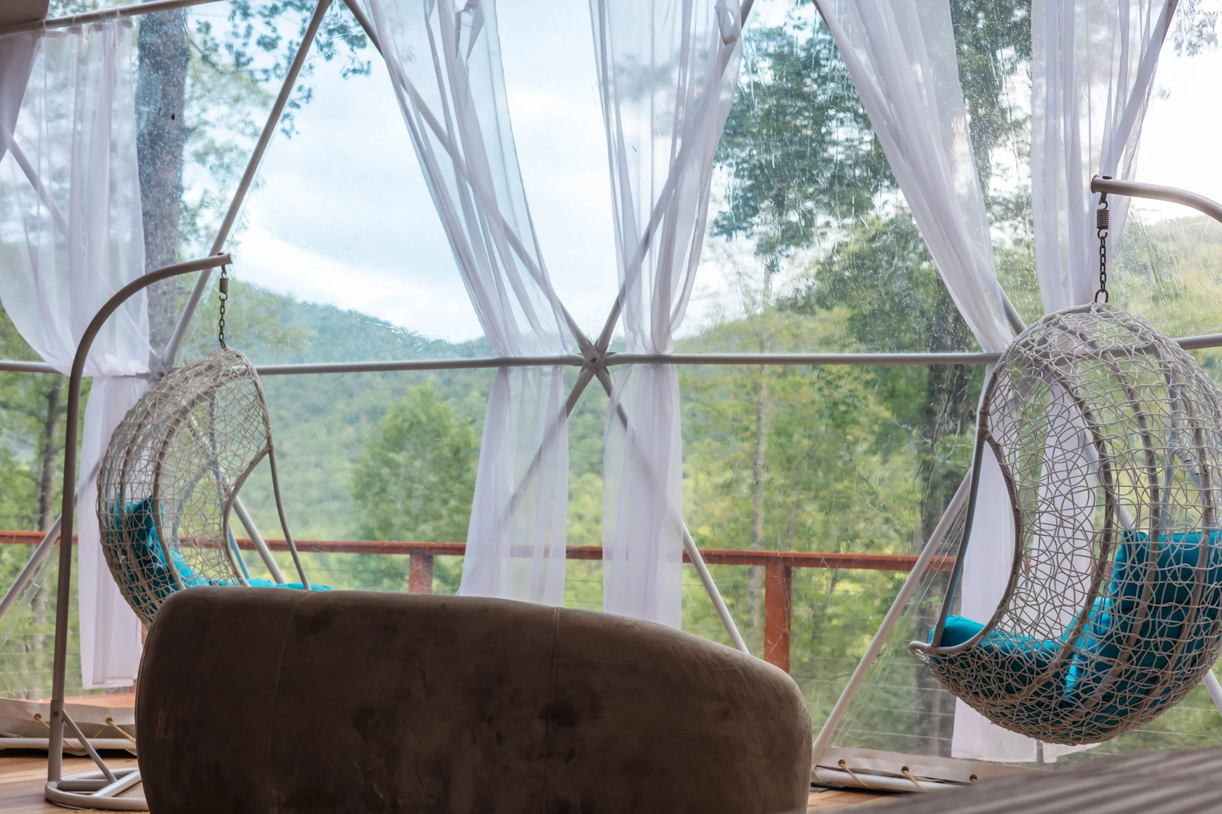 A cozy room with two hanging wicker chairs with blue cushions, a brown couch, and white curtains framing a large window overlooking green trees and a distant mountain landscape.