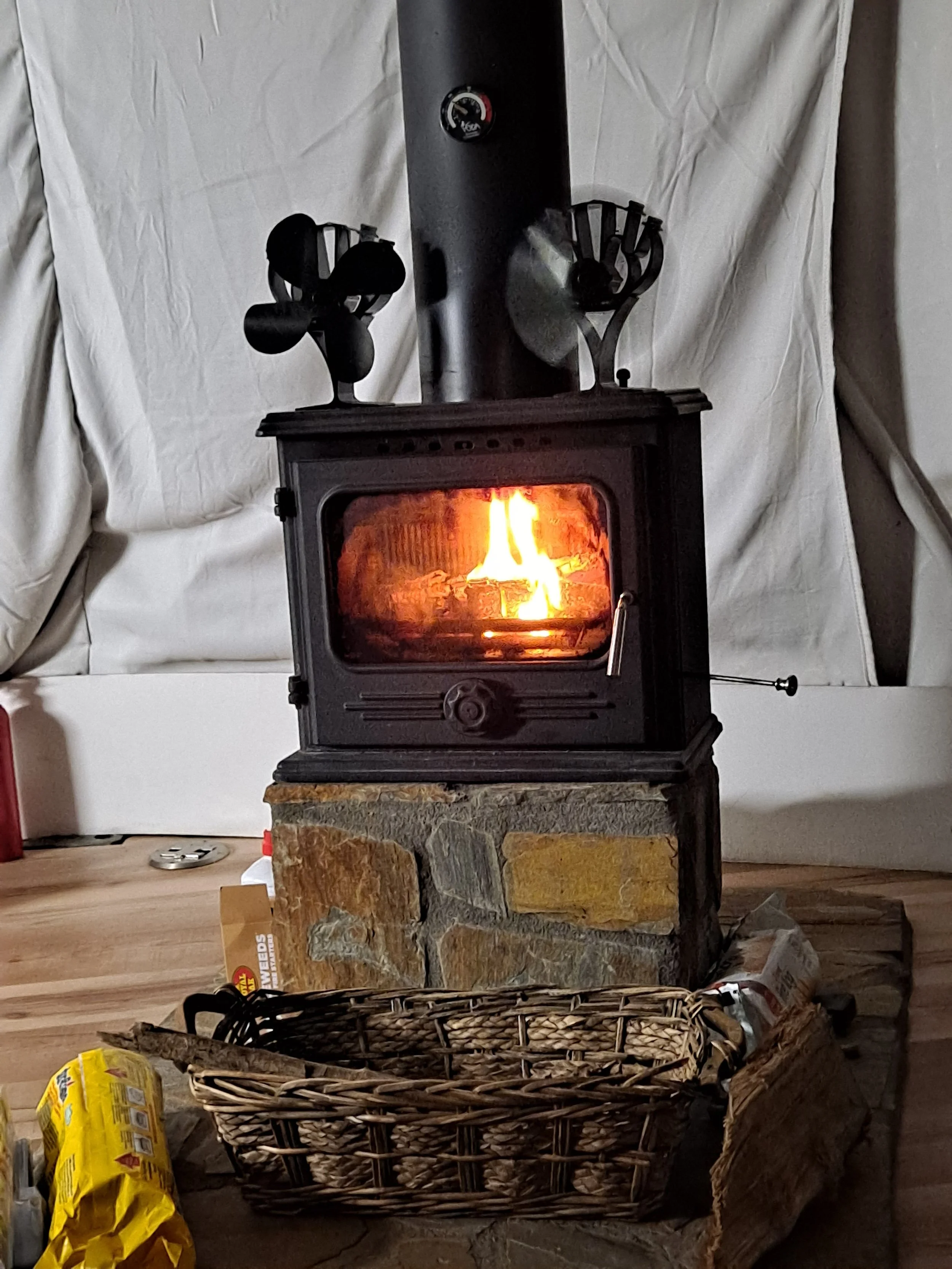 A portable stove with a fire burning inside, placed on a stone base, with a brick wall in the background and various household items around, including a wicker basket in the foreground.