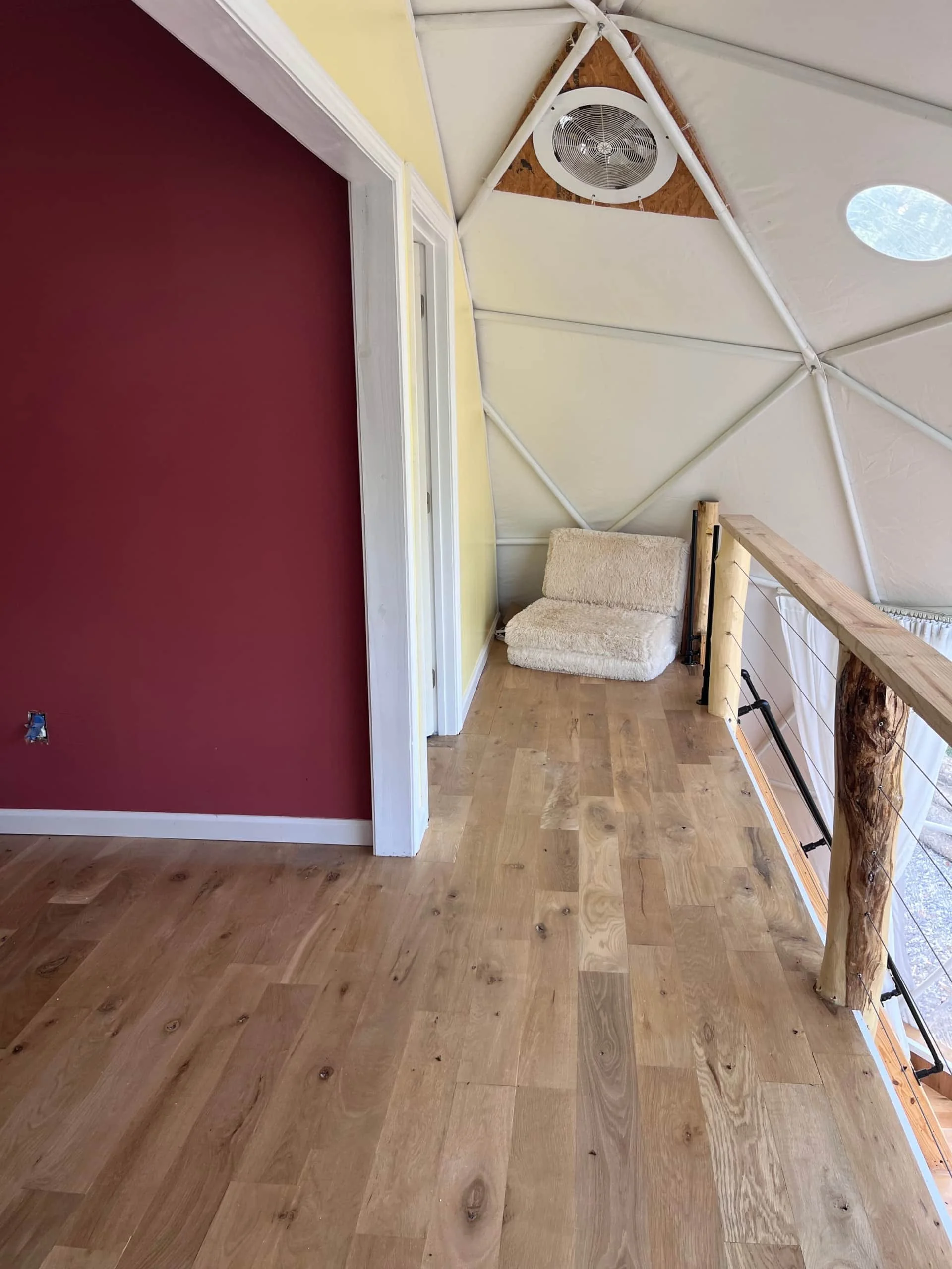 Interior of a small loft space with wooden flooring, a yellow wall, a red accent wall, a cozy beige cushion seat, and a railing with wooden logs and metal wire cables, under a white ceiling with geometric support beams and a circular window.