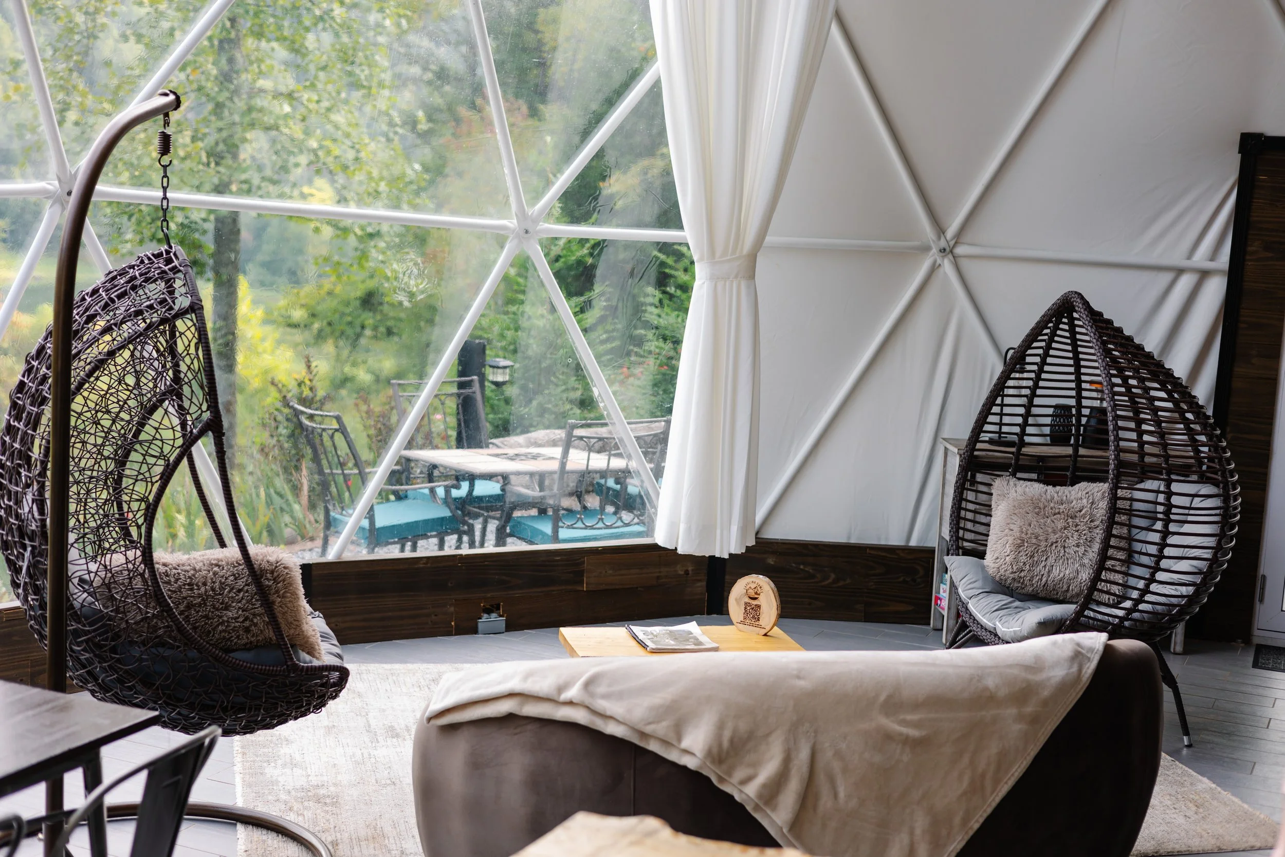Indoor seating area with two hanging wicker chairs with cushions, a small wooden table with magazines, and large geodesic window revealing a patio with outdoor table and chairs surrounded by greenery.