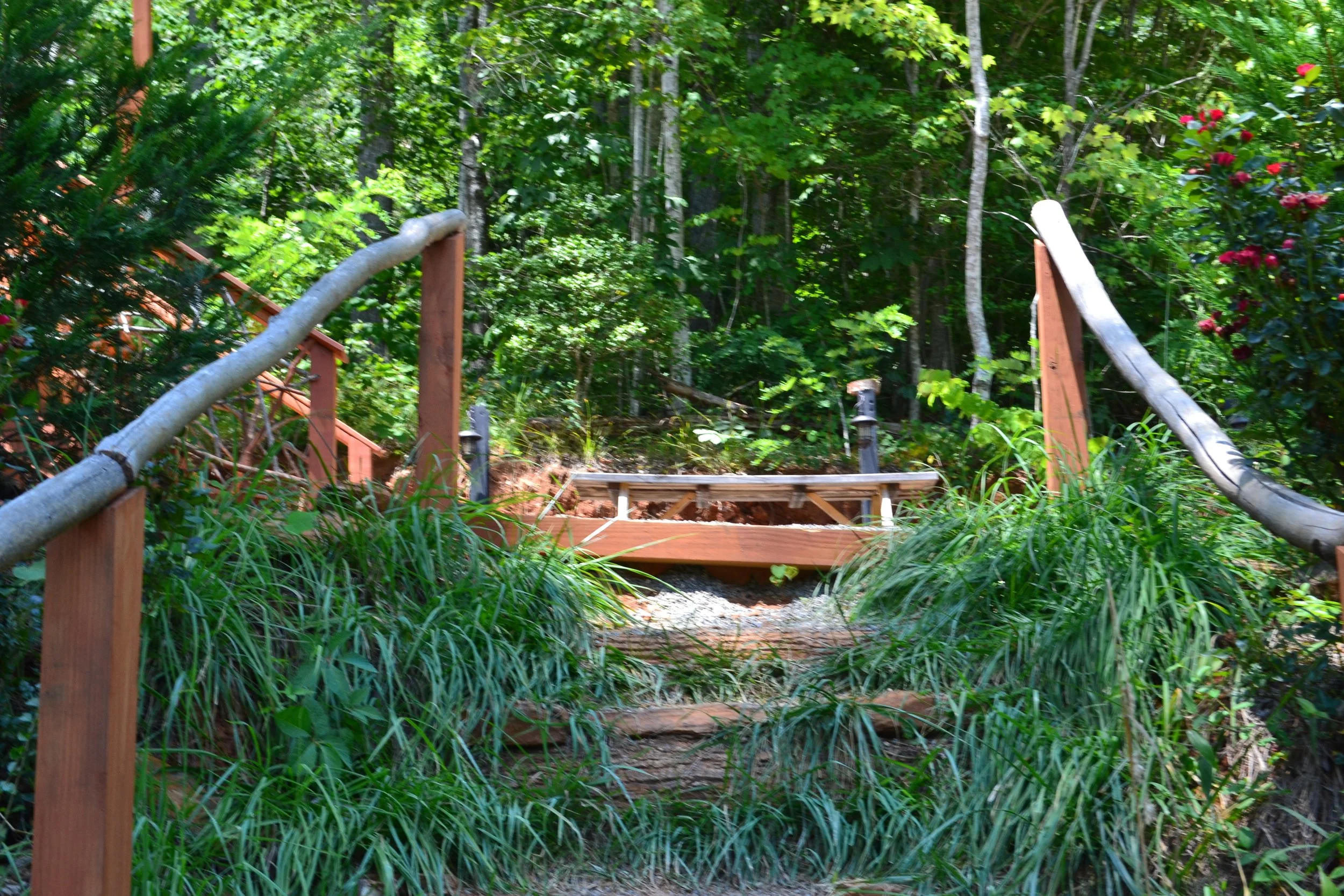 A small wooden bridge with natural log handrails crosses over a forested area with green foliage and trees on a sunny day.