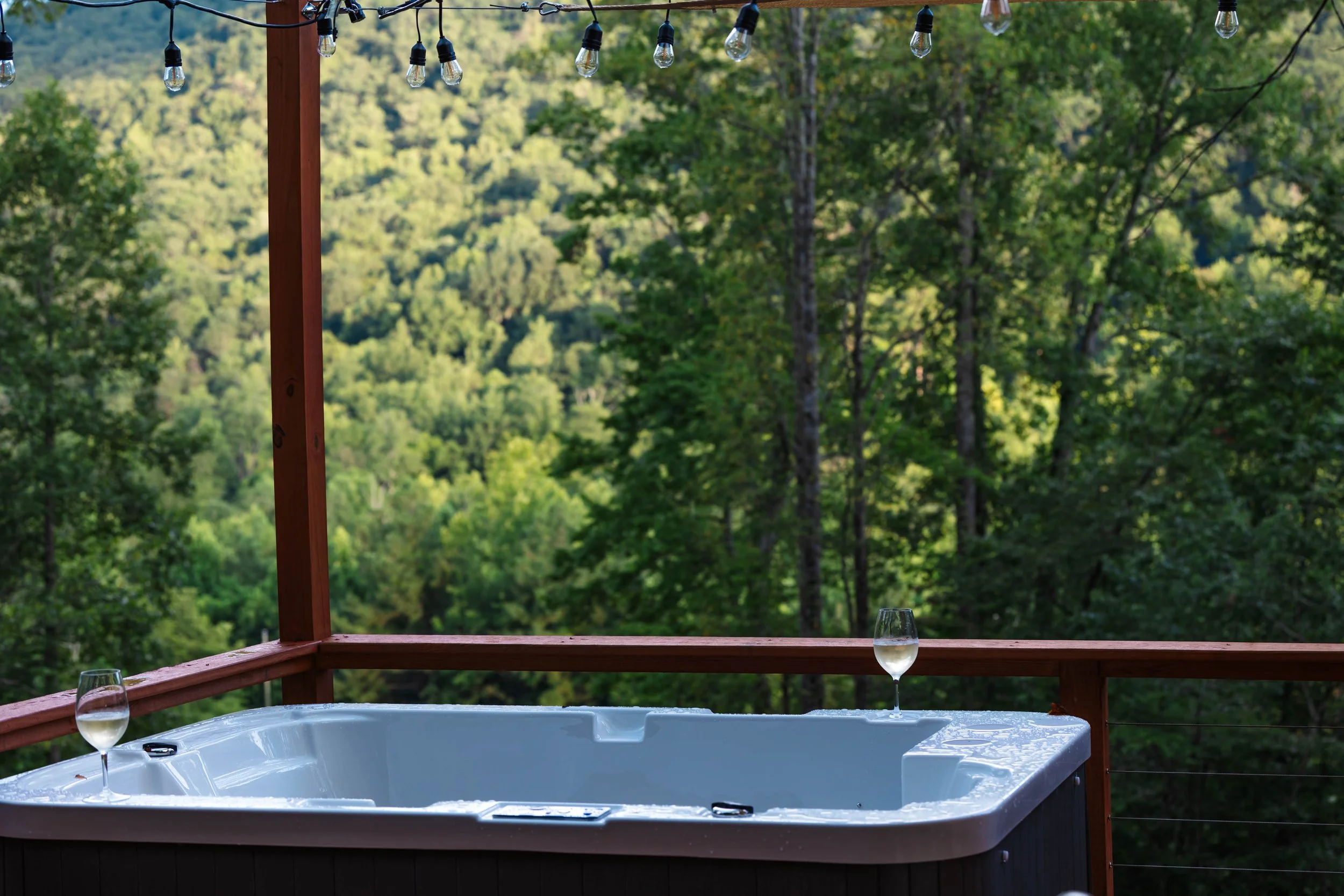 A hot tub on a wooden deck with two glasses of white wine on the edge, overlooking a lush green forest and hills.