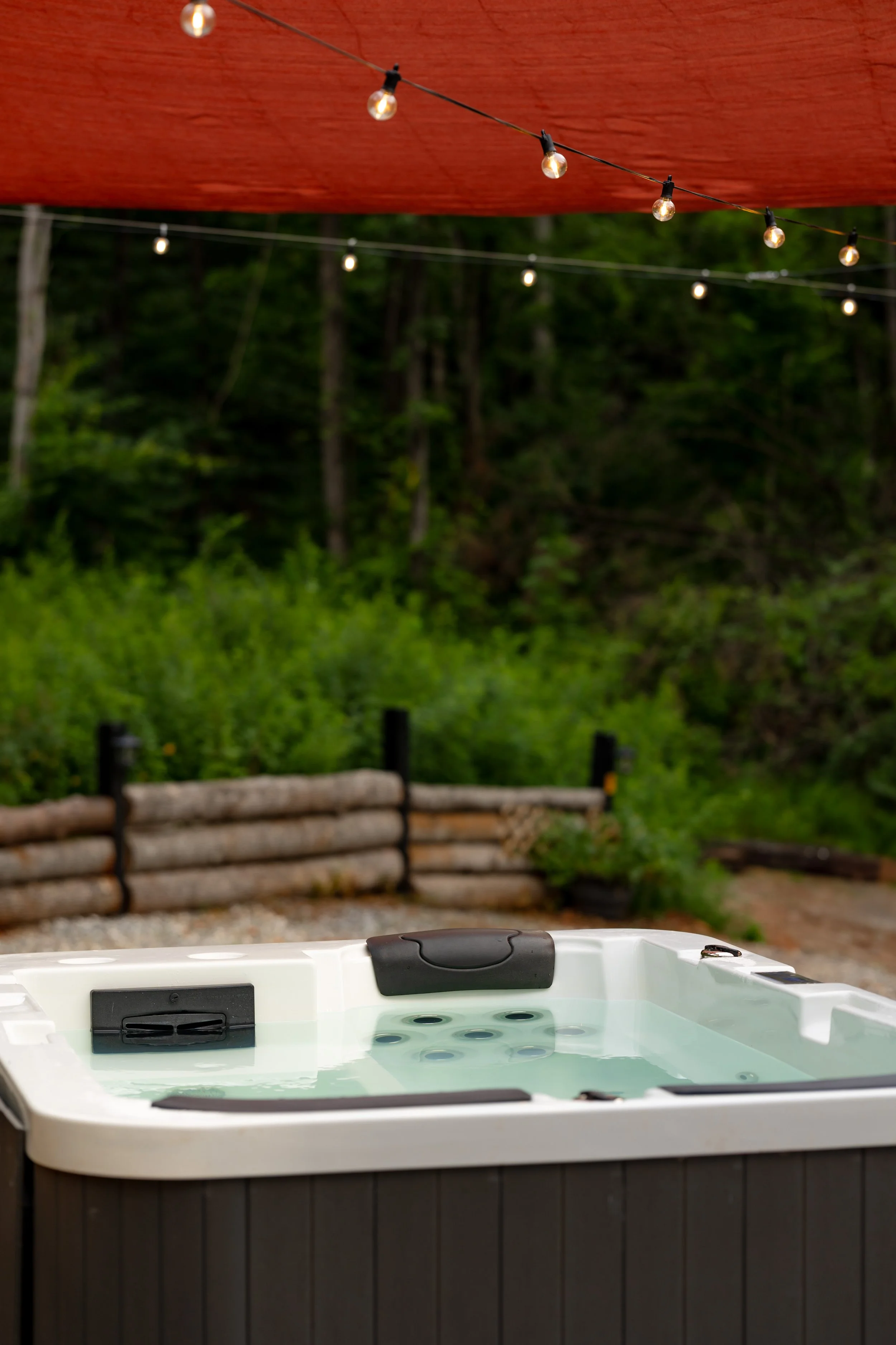An outdoor hot tub filled with water, situated under string lights, with greenery and trees in the background.
