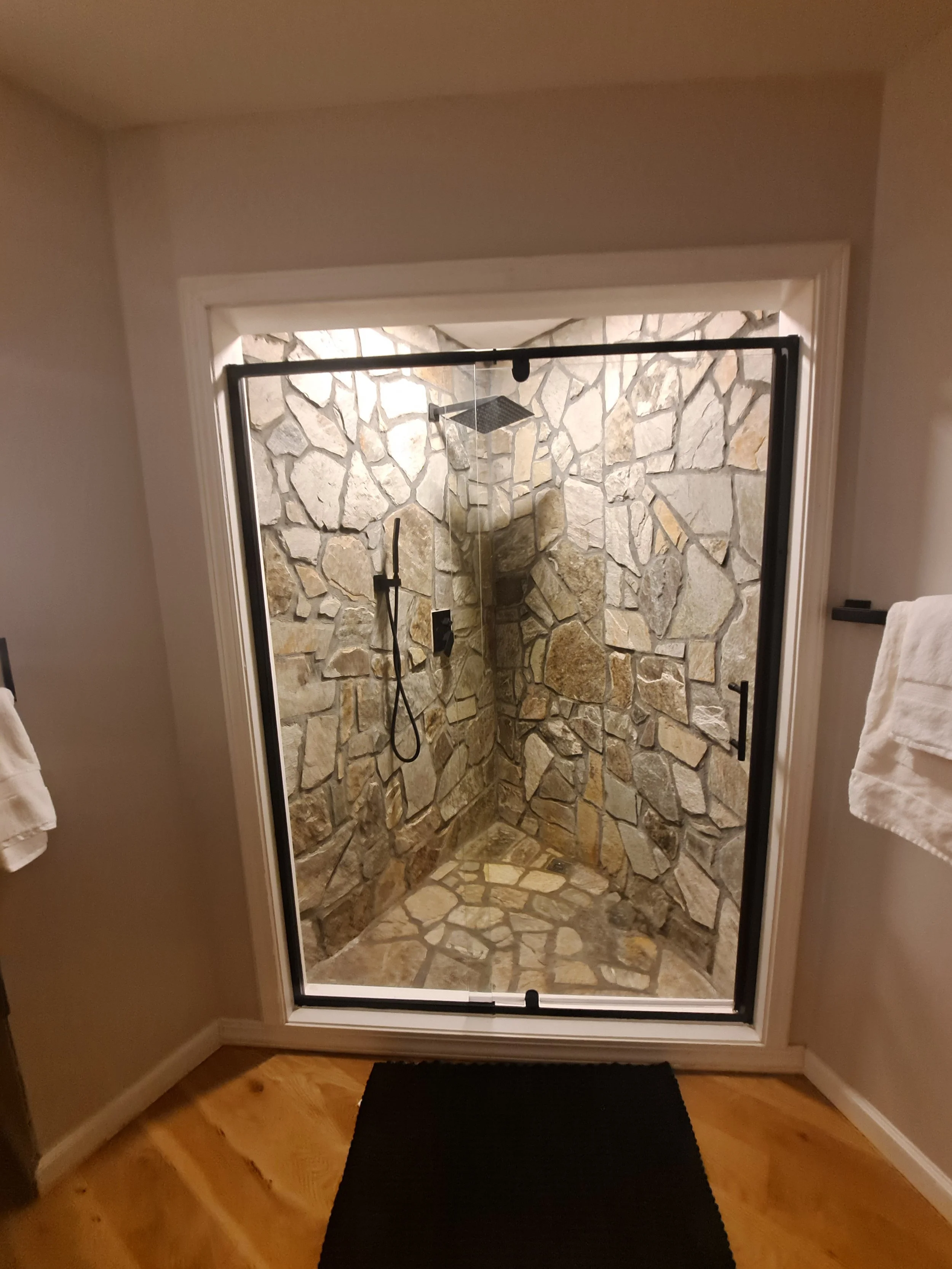 Indoor walk-in shower with stone wall, glass door, and rainfall showerhead.