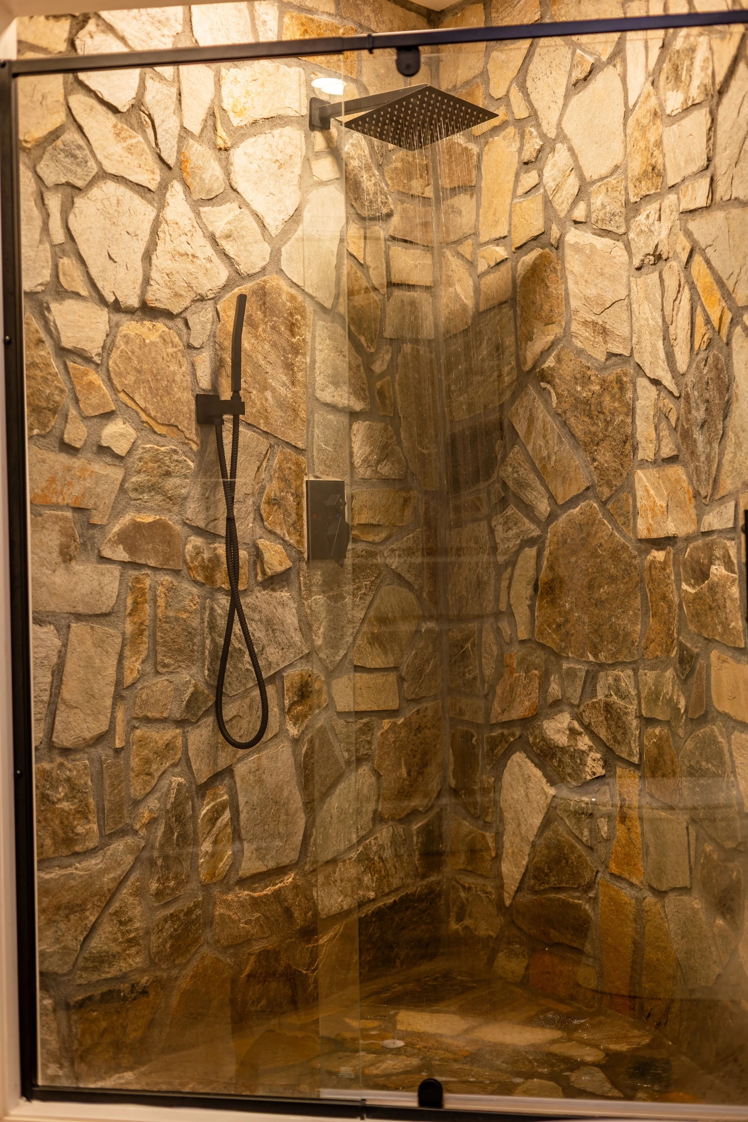 A walk-in shower with stone tile walls, a large square showerhead, a handheld shower wand, and a glass door.