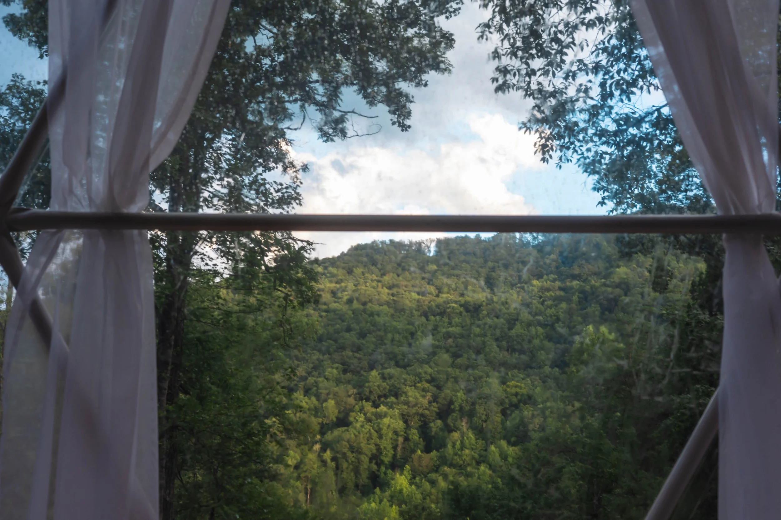View through a window with white curtains showing a lush green forested hillside under a partly cloudy sky.