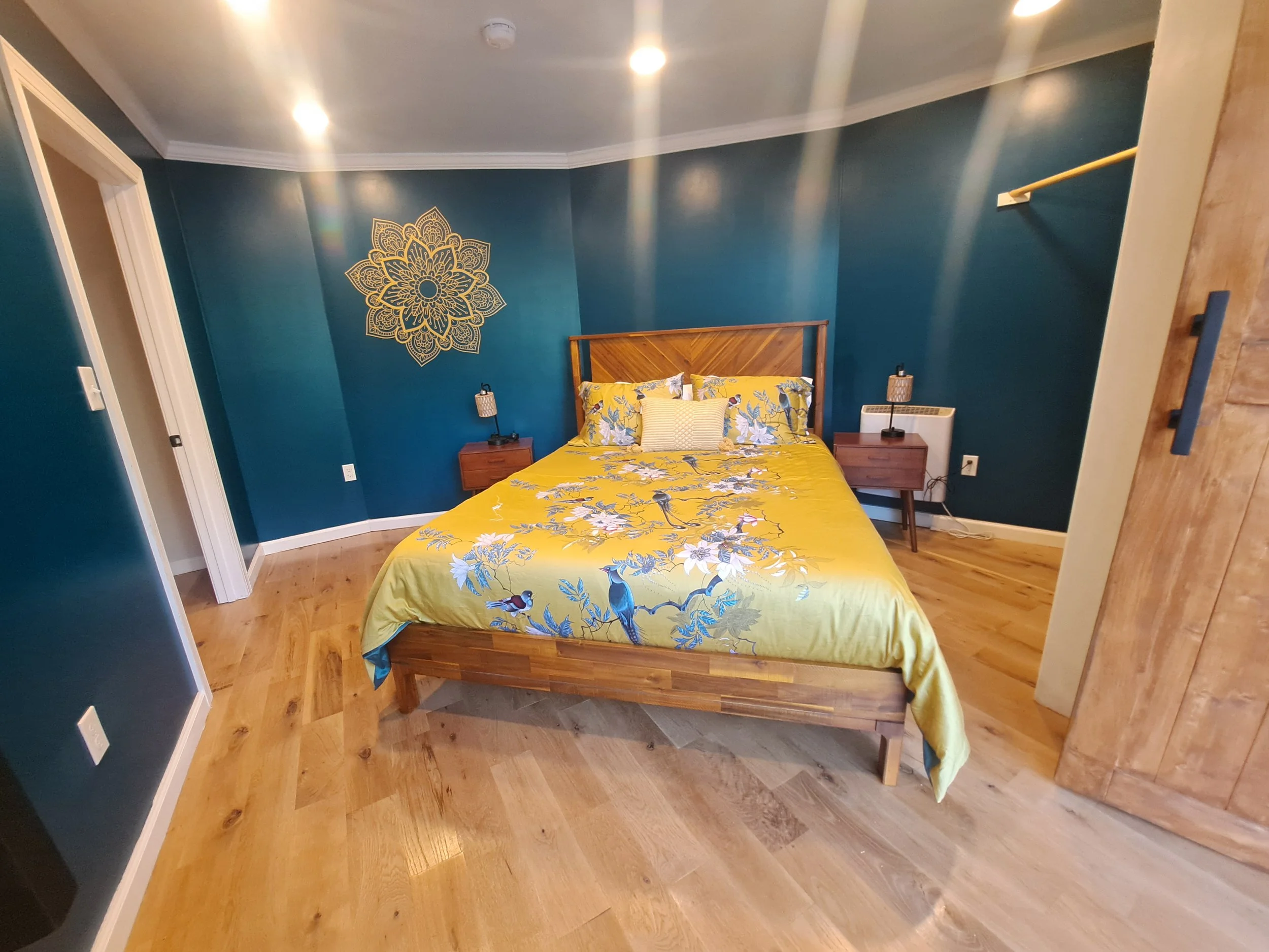 A bedroom with dark teal walls, a wooden headboard, and yellow bedding with a bird and floral pattern. There are two pillowy nightstands with lamps on each side of the bed. The floor is light-colored hardwood, and there is a decorative gold mandala w