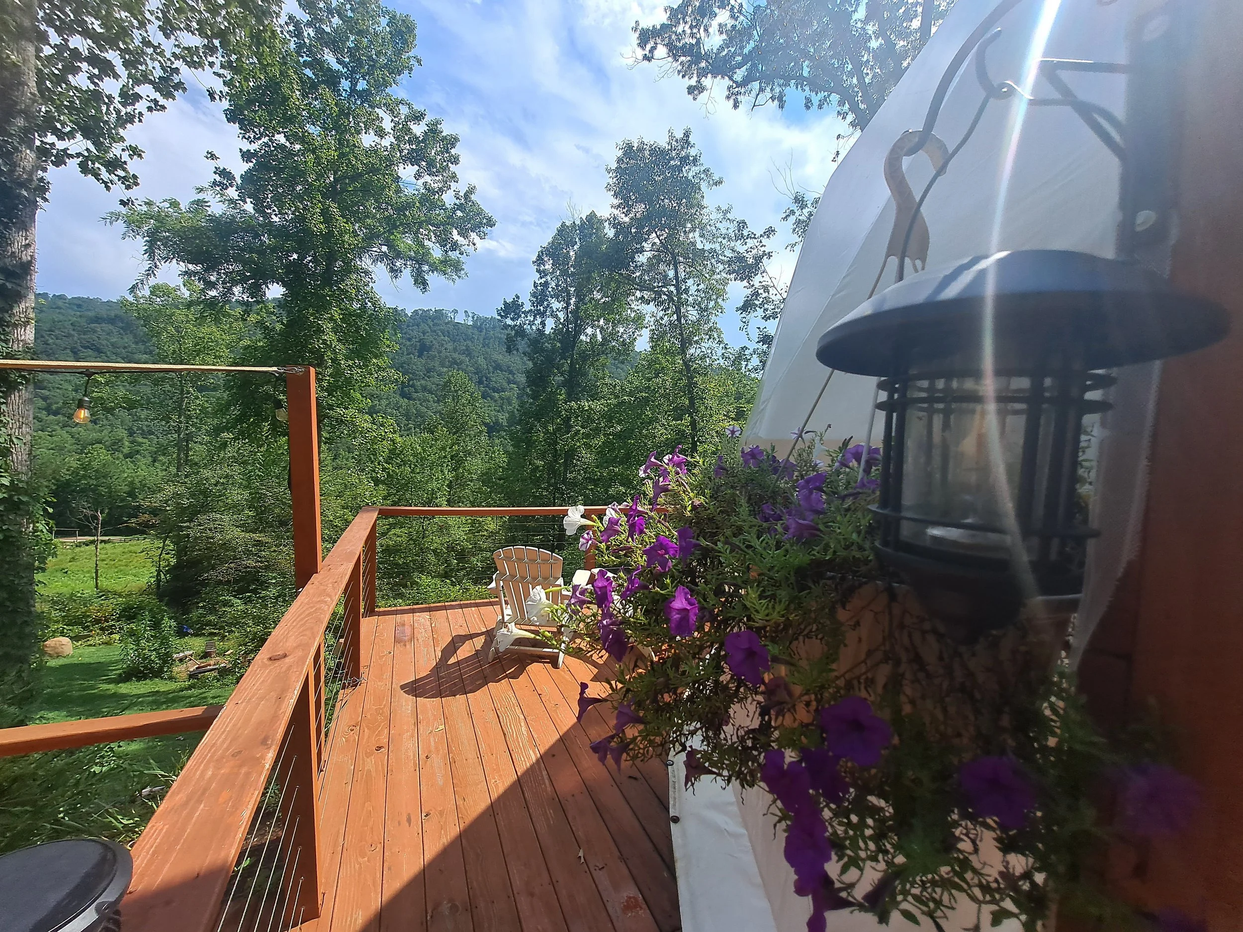 Wooden deck with purple flowers in a hanging basket, a white Adirondack chair, trees, mountains, and a partly cloudy sky.