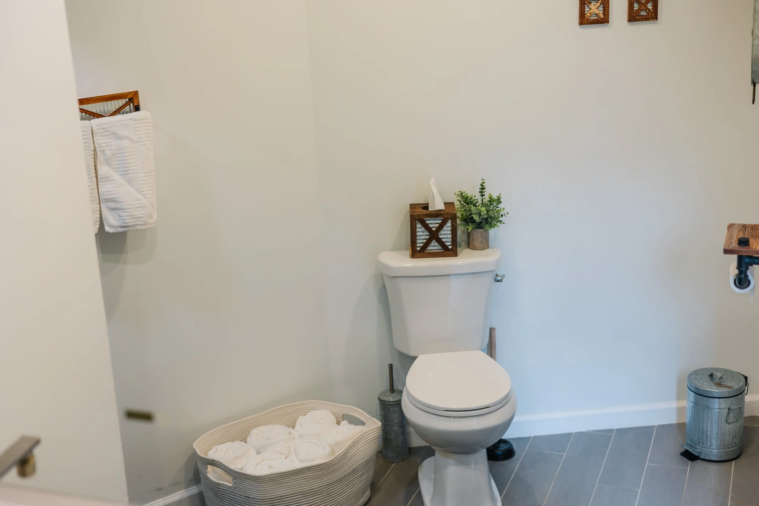 A bathroom featuring a white toilet with a decorative box and plant on top, a basket of rolled towels on the floor, a trash can, a towel rack with white towels, and wooden wall decorations.