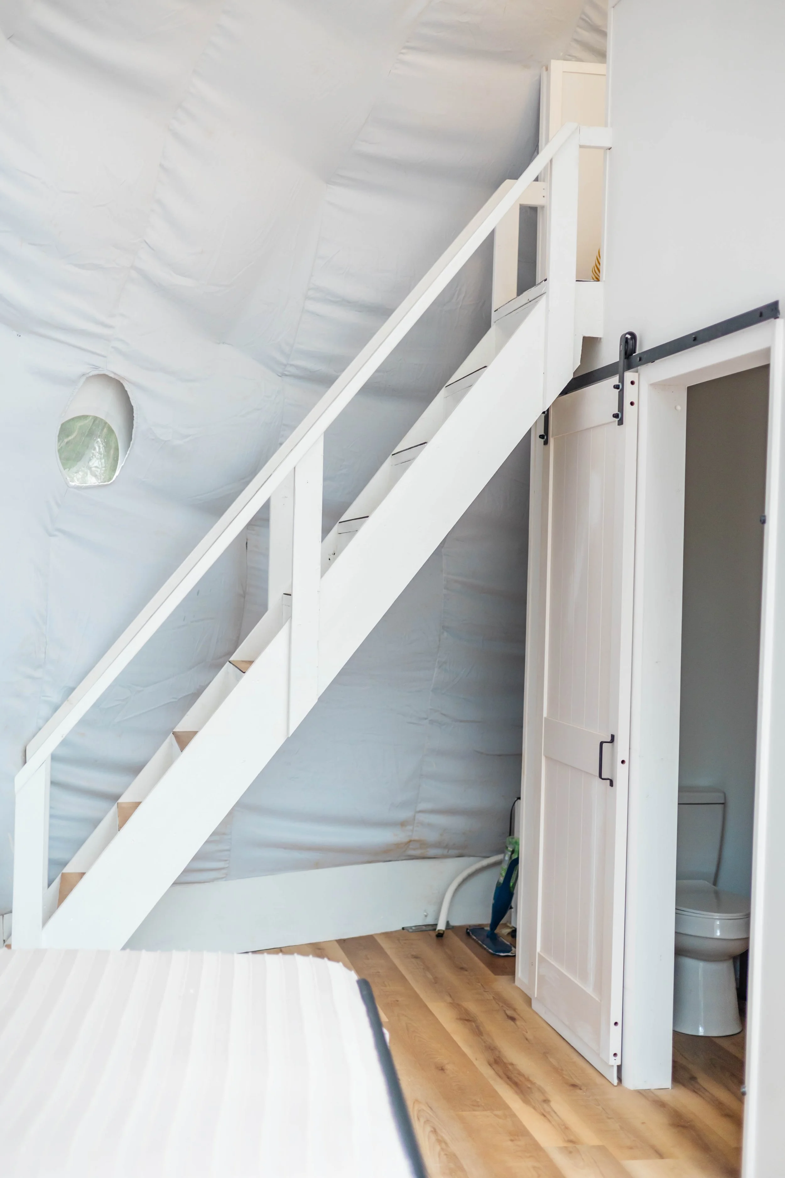 Interior of a room under construction or renovation with white staircase leading to a loft or upper level, small bathroom with a toilet behind a white door, wood flooring, and unfinished insulation on the wall.