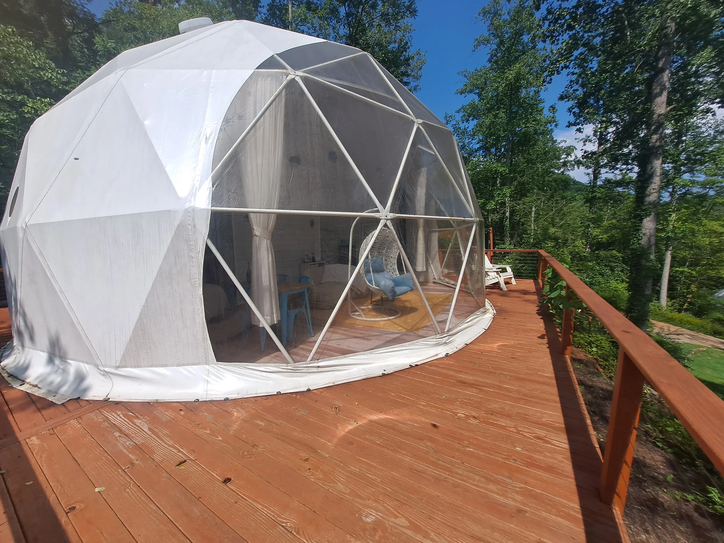 A backyard wooden deck with a clear geodesic dome tent surrounded by green trees. Inside the tent, there's a small sofa and a blue chair visible through the transparent walls.
