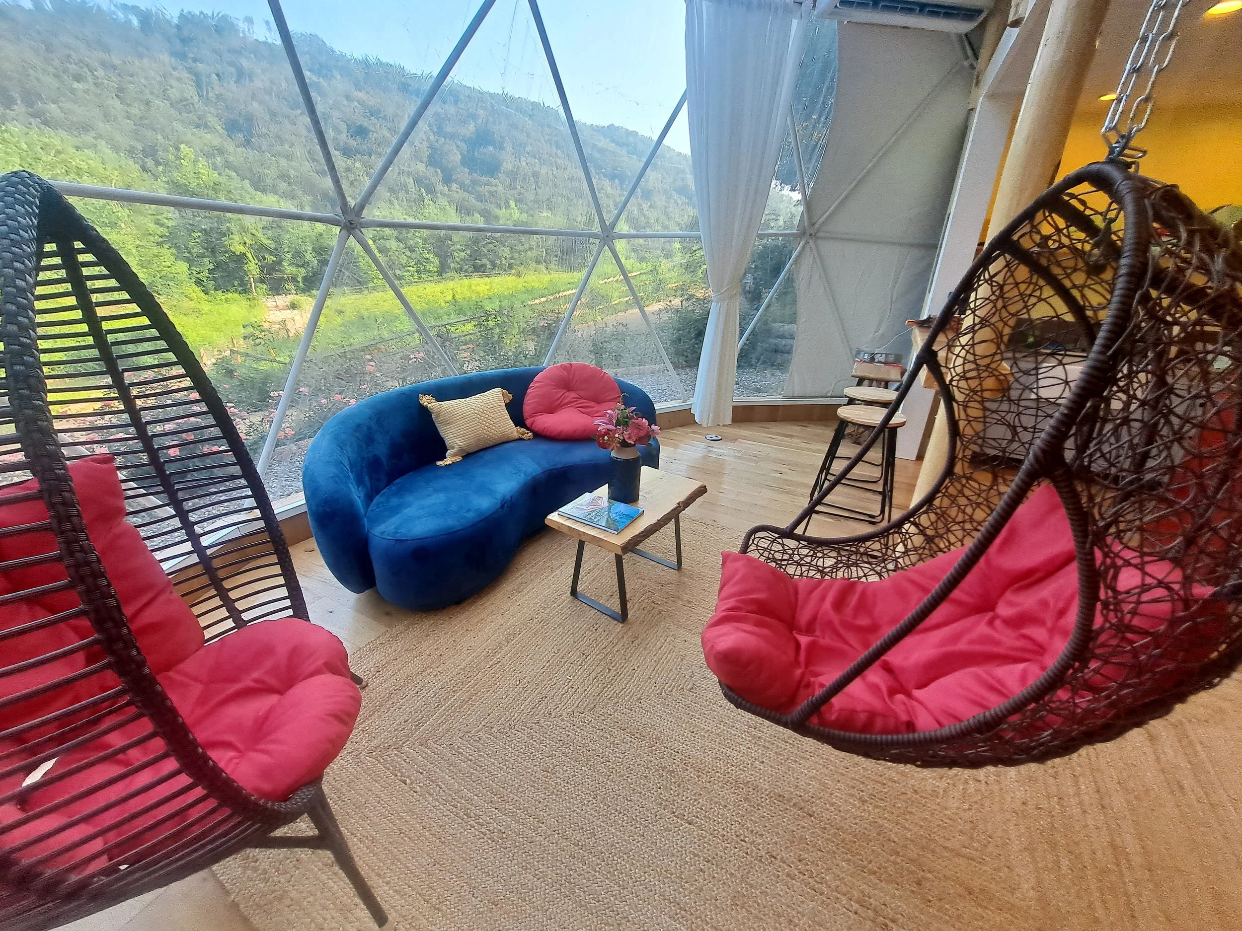 Indoor seating area with large window view of green mountains, featuring two hanging chairs with pink cushions, a blue velvet sofa with colorful pillows, a small table with flowers, and four chairs in the background.
