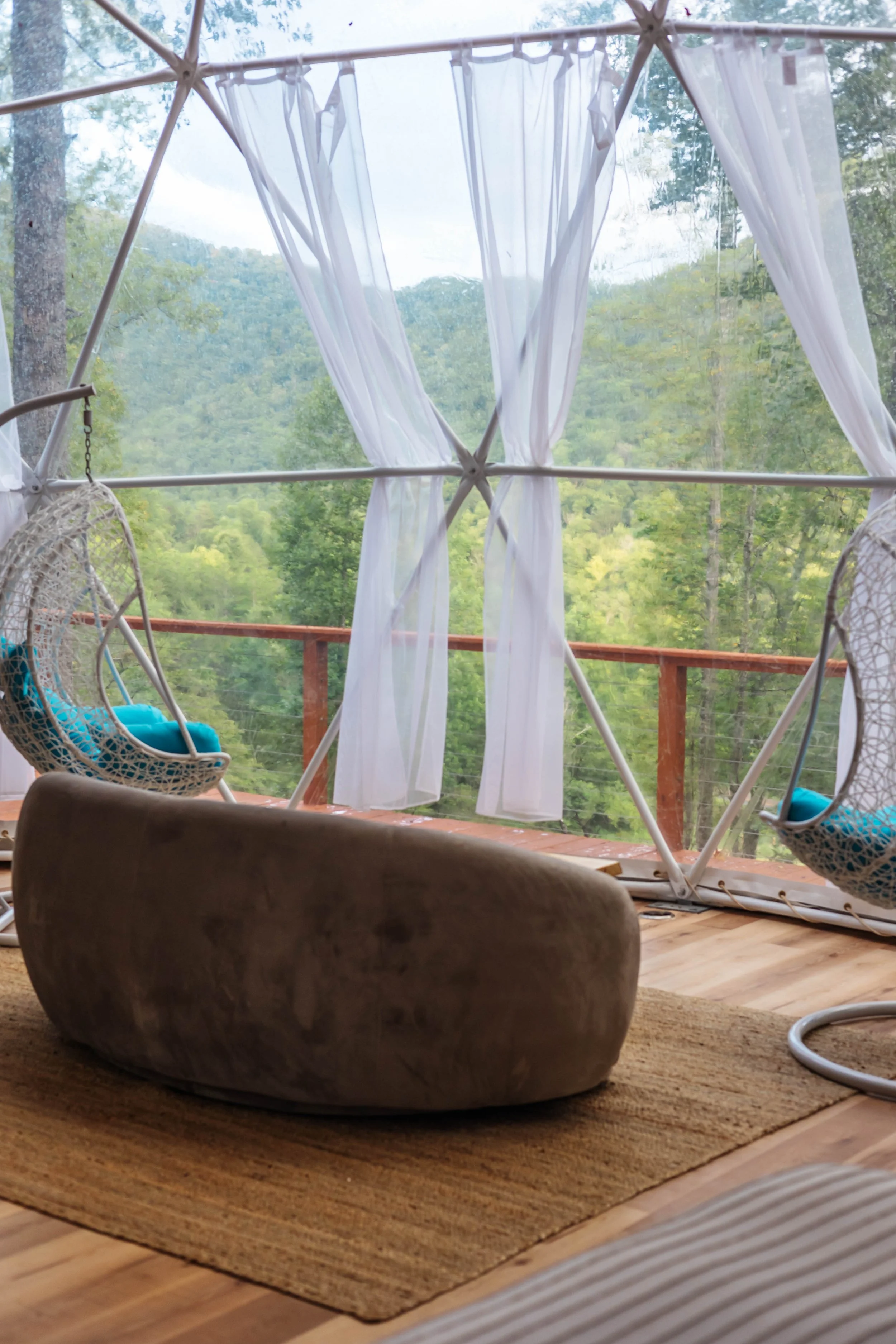 A cozy indoor space with large glass windows revealing a lush green forest and mountains outside. Inside, there are two hanging rattan chairs with blue cushions, a smooth stone ottoman on a woven rug, and white curtains tied back at the windows.