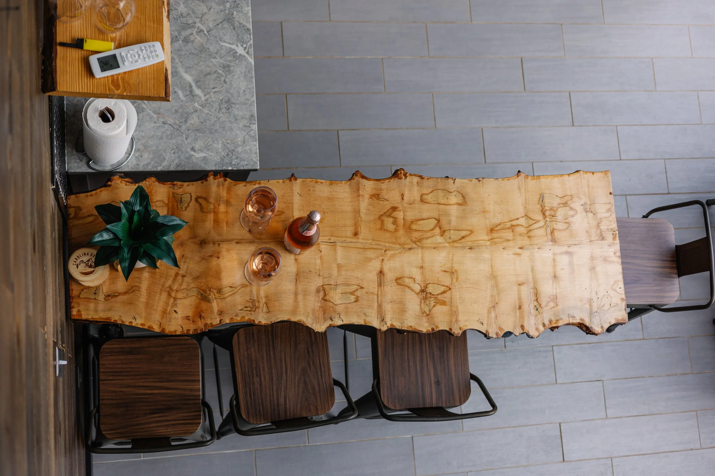 A rustic wooden dining table with a live edge, set with two glasses of rosé wine and a bottle of rosé, surrounded by four dark wood chairs with metal frames, in a modern interior with gray tiled flooring and a marble counter with a paper towel roll a