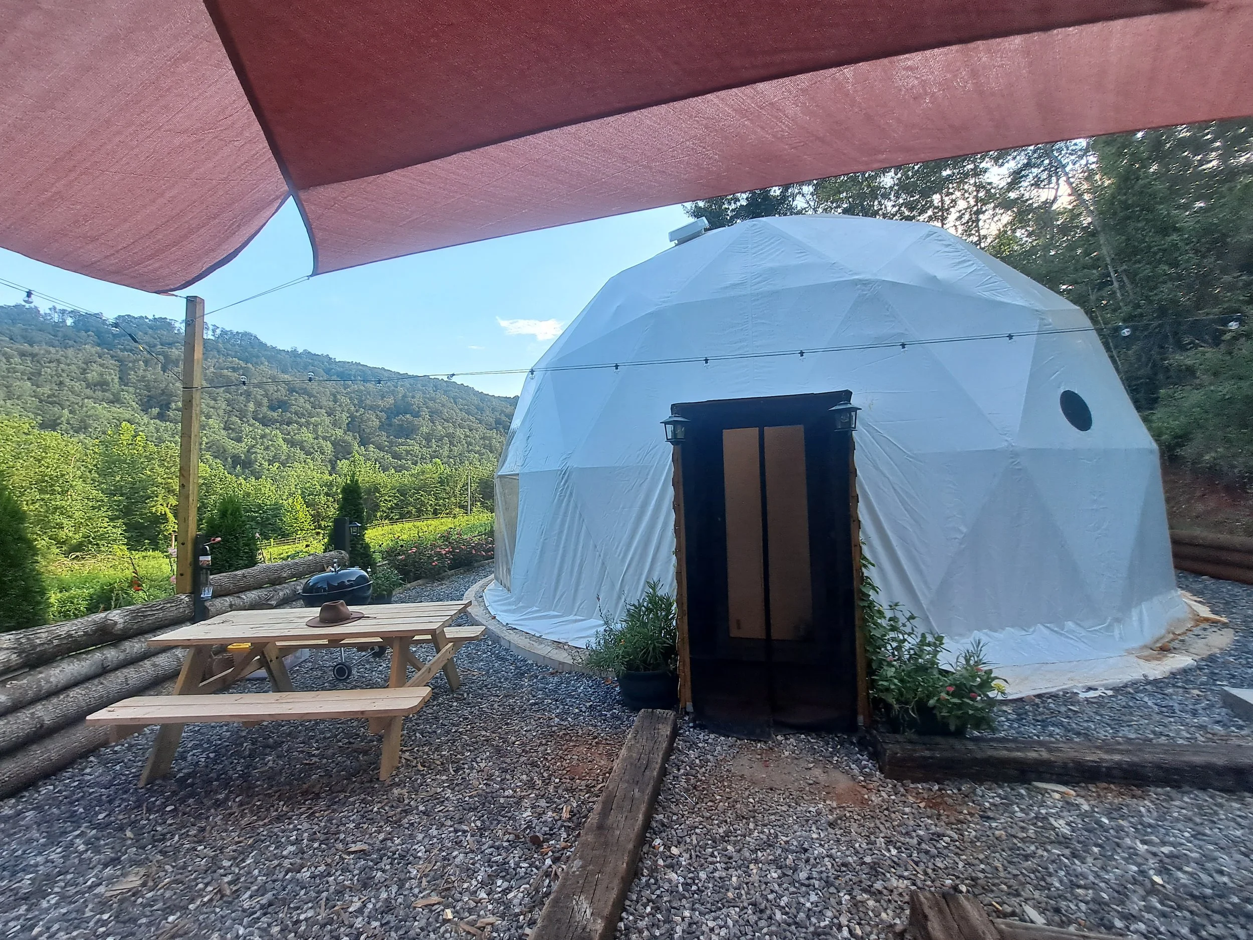 A geodesic dome tent outdoors on a gravel area, with a picnic table and a hat on top, surrounded by trees and hills under a blue sky.