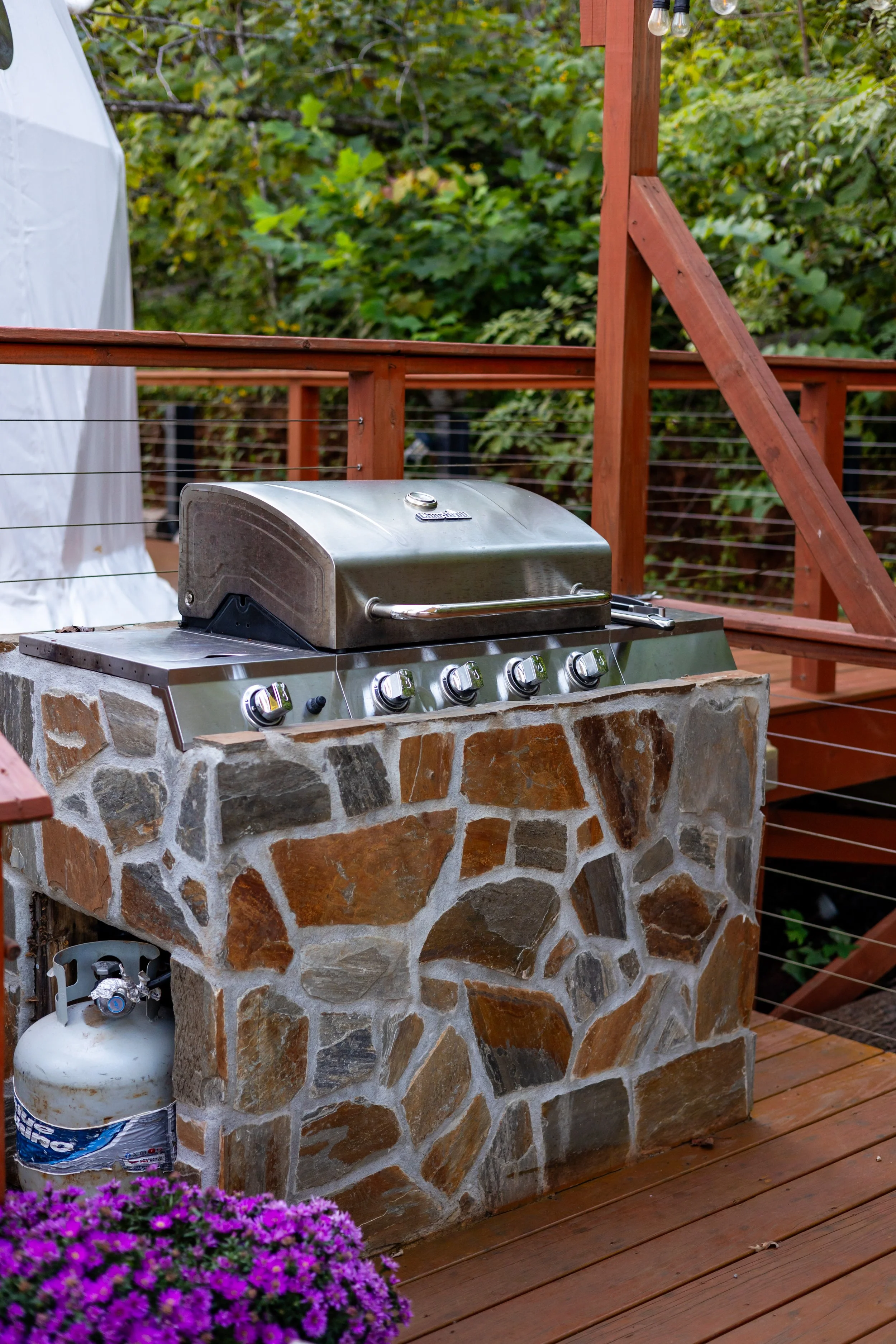 A stainless steel outdoor gas grill with five control knobs, built into a stone counter on a wooden deck, with a propane tank underneath, and purple flowers in a pot nearby.