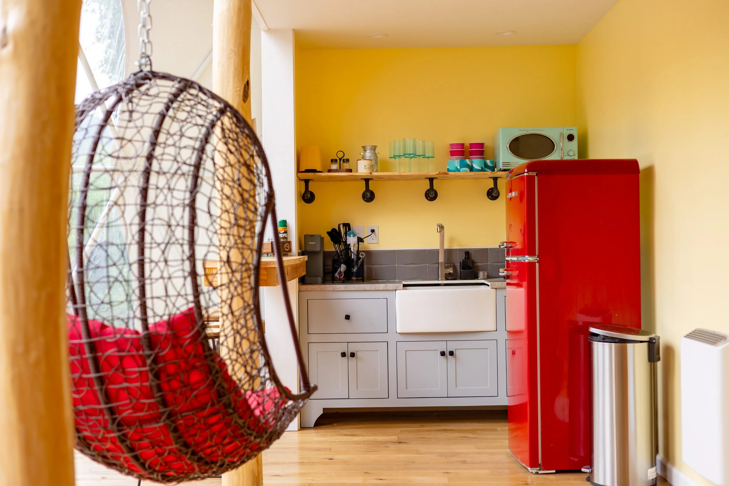 Colorful kitchen with yellow walls, red refrigerator, and gray cabinetry; hanging birdcage with red balls in foreground.