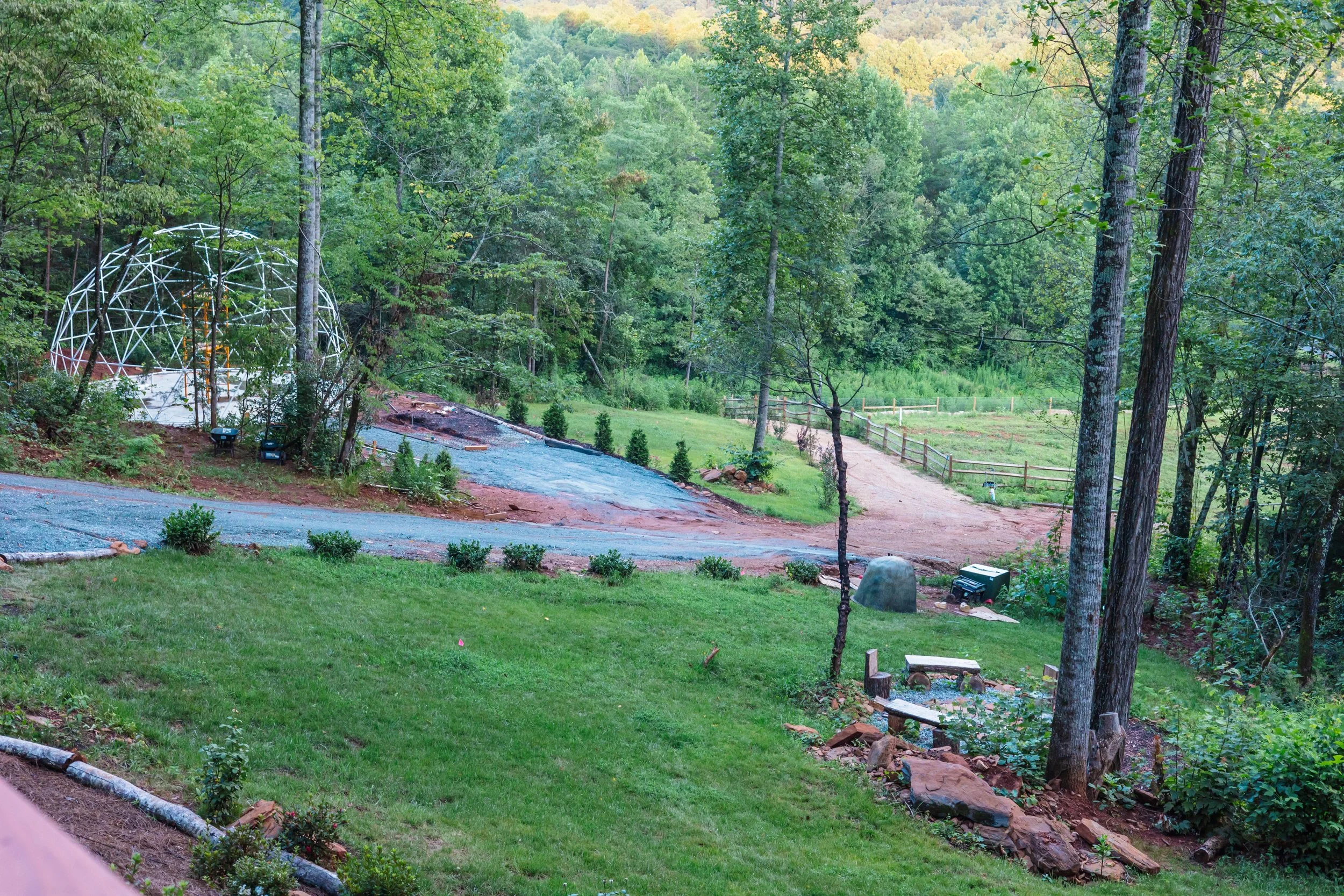 A backyard with a grassy lawn, small shrubs, and trees. There is a partially assembled geodesic dome structure on the left side. A dirt pathway leads into a dense forested area in the background.