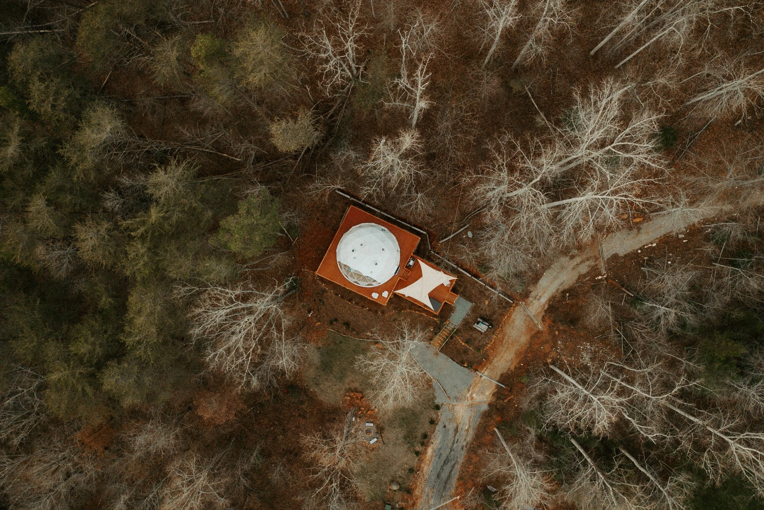 An aerial view of a house with a red roof and a white rooftop deck in a wooded area during fall, with trees and a winding dirt road surrounding the property.