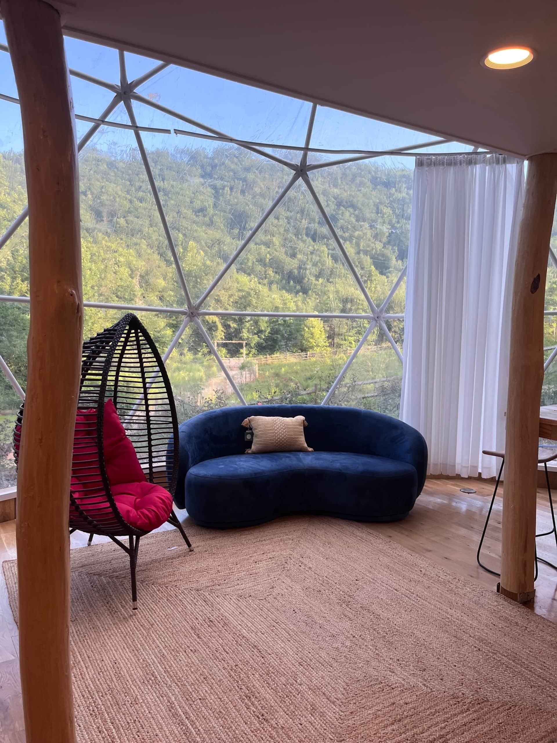 Interior of a glamping dome with a large window overlooking green hills, a blue velvet sofa with a beige pillow, a hanging black egg chair with a pink cushion, a beige rug, and white curtains.