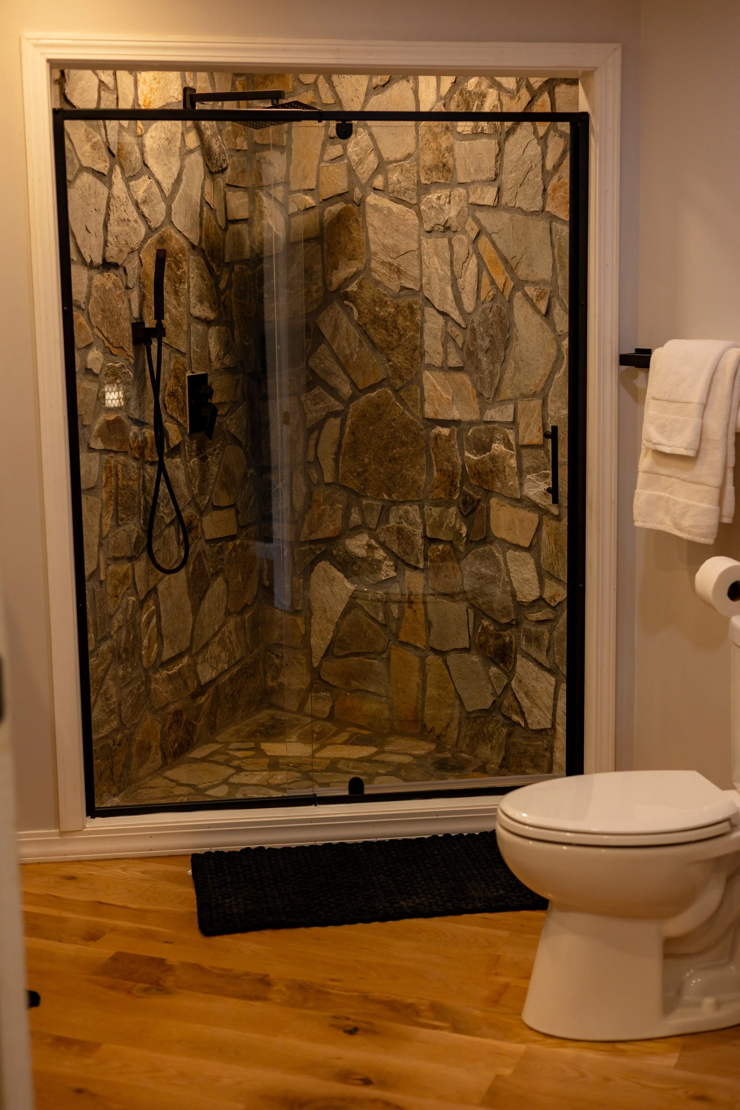 A bathroom with a stone shower, a toilet, a towel rack with a white towel, and a black bath mat on a wooden floor.