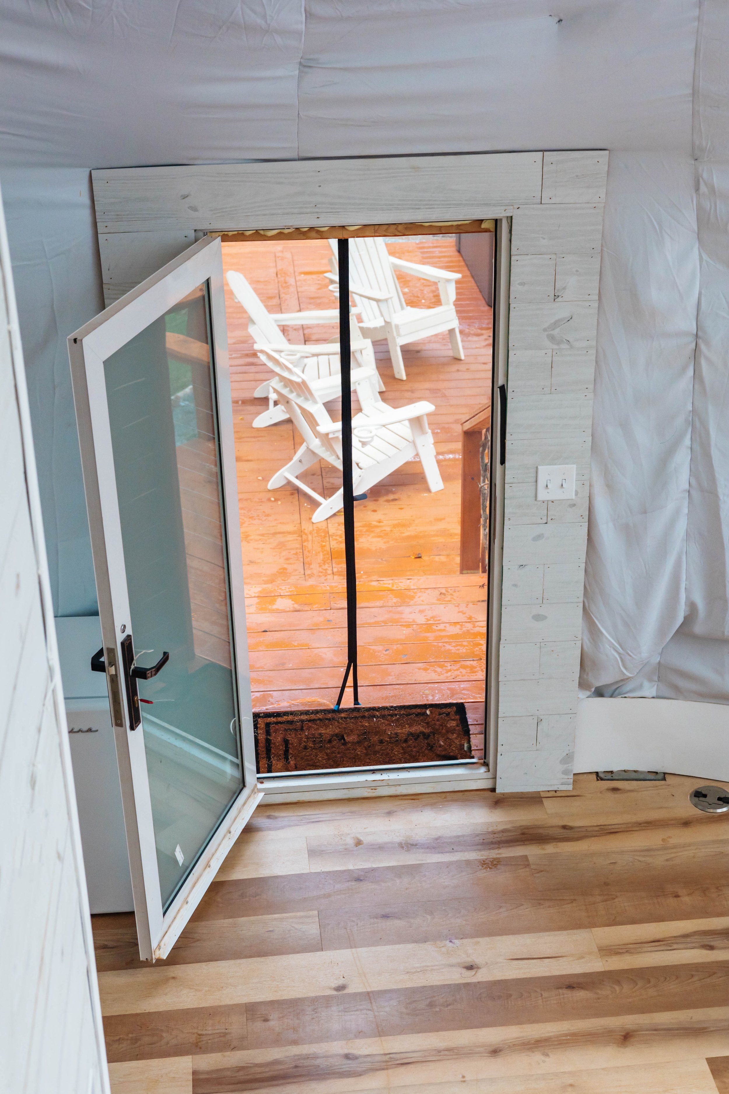 View of a glass door leading to an outdoor wooden deck with white chairs. The indoor area has wooden flooring and white walls.