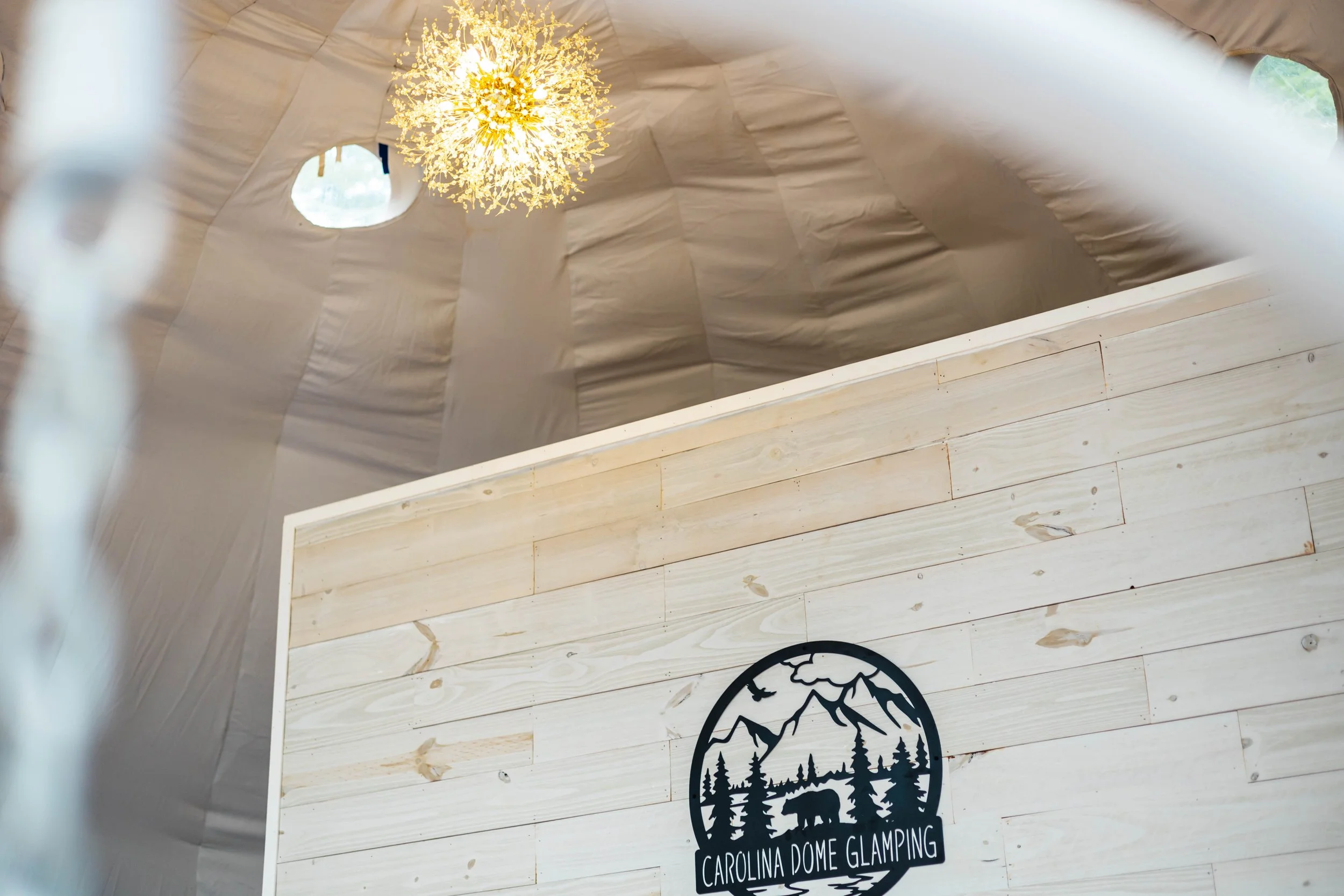 Interior of a glamping dome with a wooden wall and a logo for Carolina Dome Glamping featuring mountains, trees, and a bear, along with a star-shaped chandelier hanging from the ceiling.