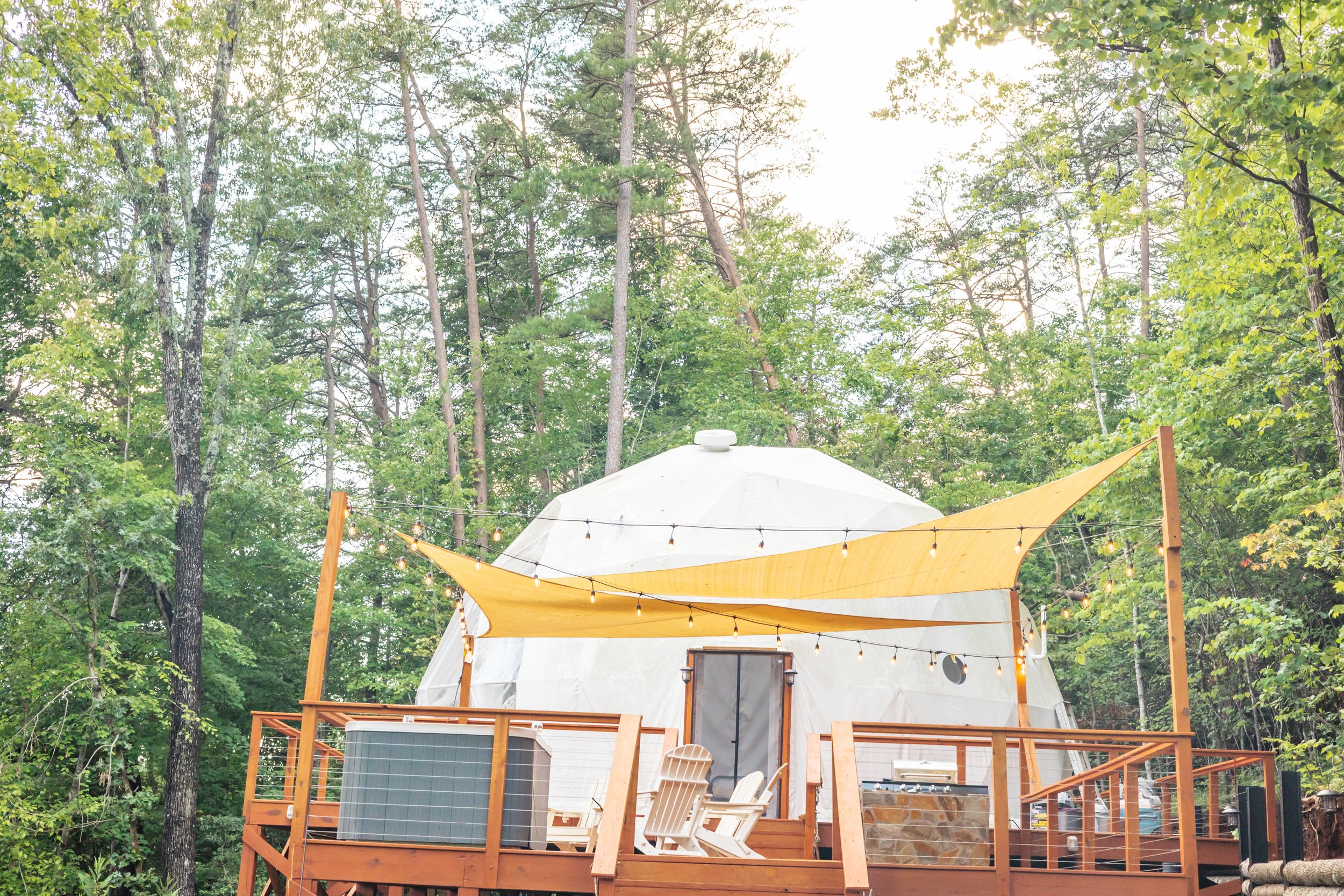 A treehouse with a white dome-shaped roof, surrounded by green trees. The treehouse features a wooden deck with Adirondack chairs, string lights, and a yellow canopy for shade.