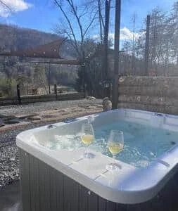 Hot tub outdoors with two glasses of champagne floating on the water, surrounded by a wooden deck and a wooded area with trees.