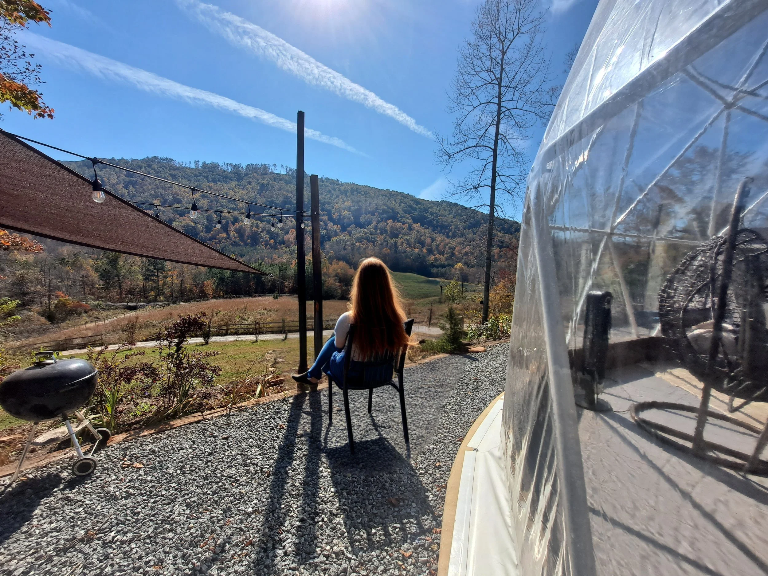 A woman with long hair sitting on a chair outdoors, facing a scenic mountain landscape with clear blue skies.