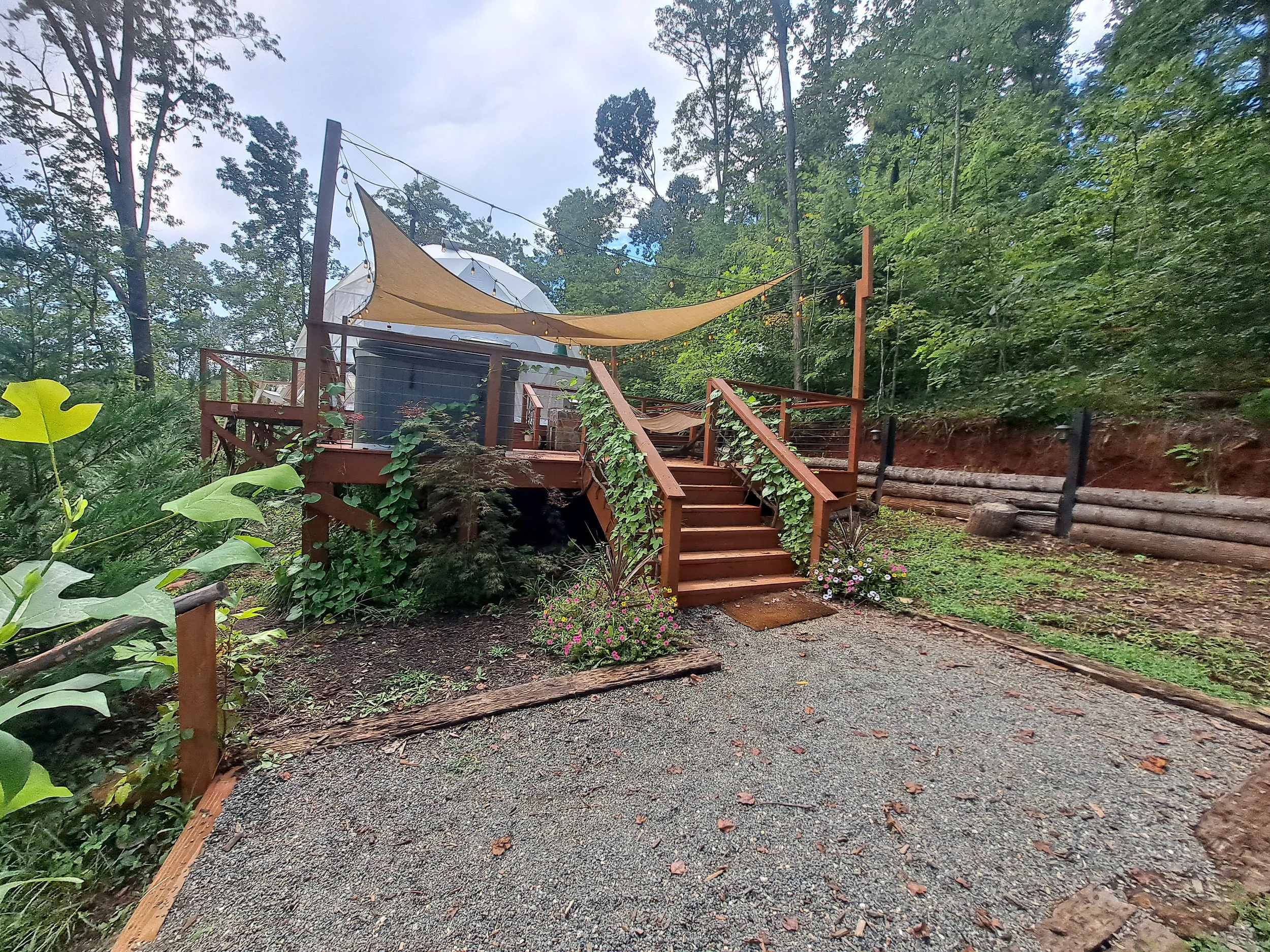 Wooden deck with steps leading up to a boat-shaped seating area, decorated with string lights and beige shade sail, surrounded by greenery and trees.