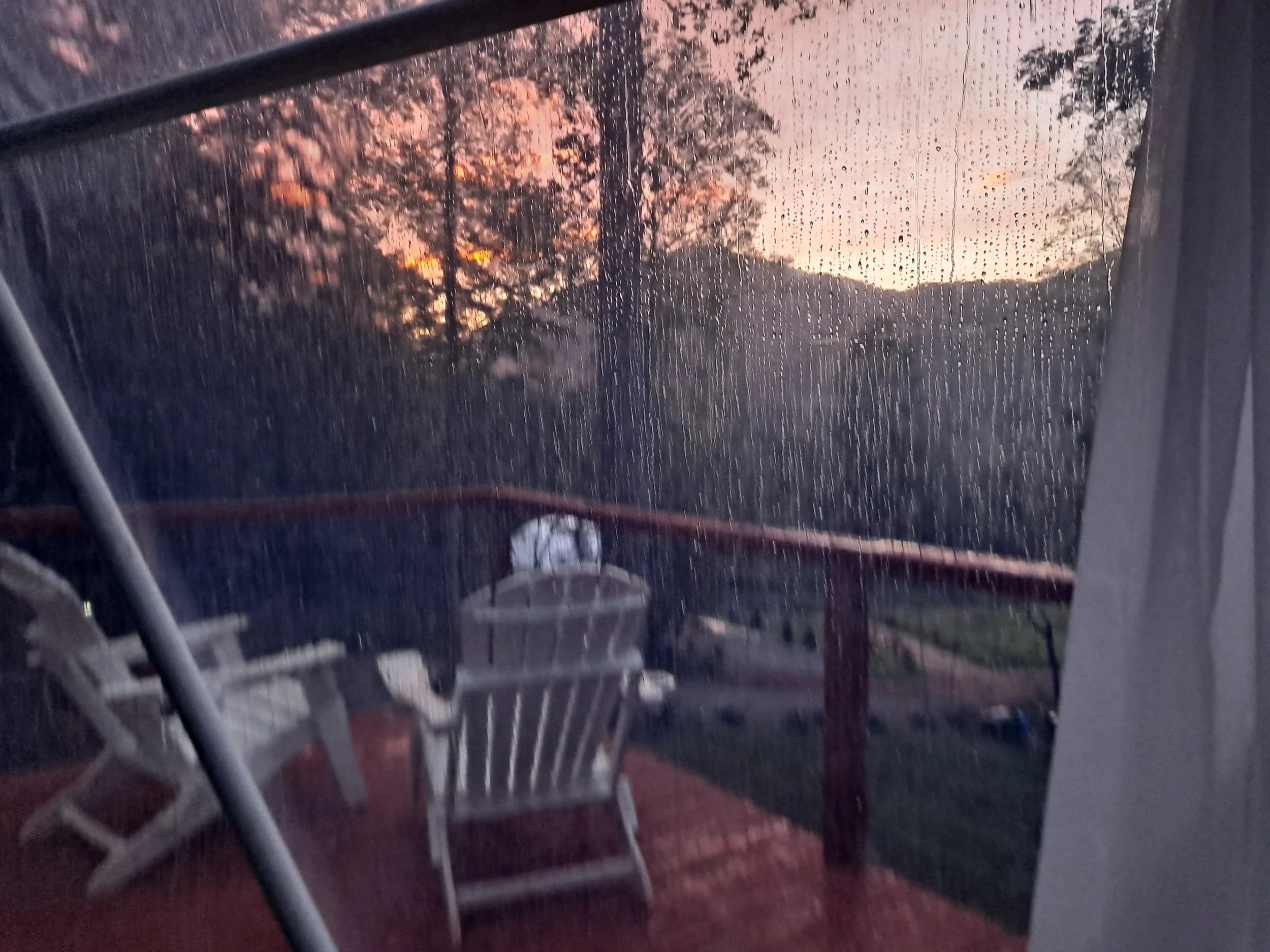 Rain on a window with a view of a wooden deck, plastic chairs, and trees at sunset