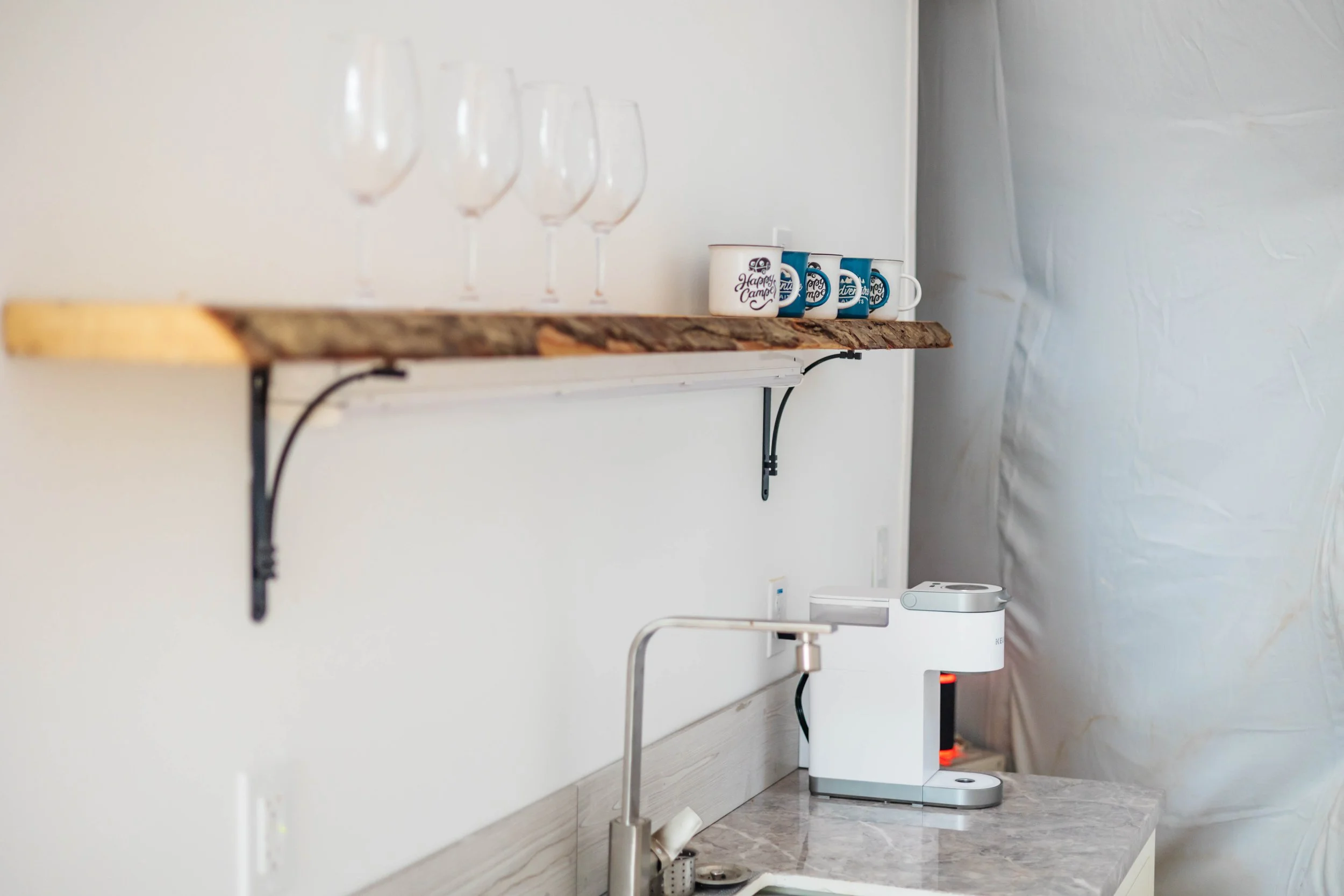 Kitchen counter with a marble surface, a white coffee machine, a stainless steel sink, a wooden shelf holding three wine glasses, some coffee mugs, and a wall-mounted light fixture.