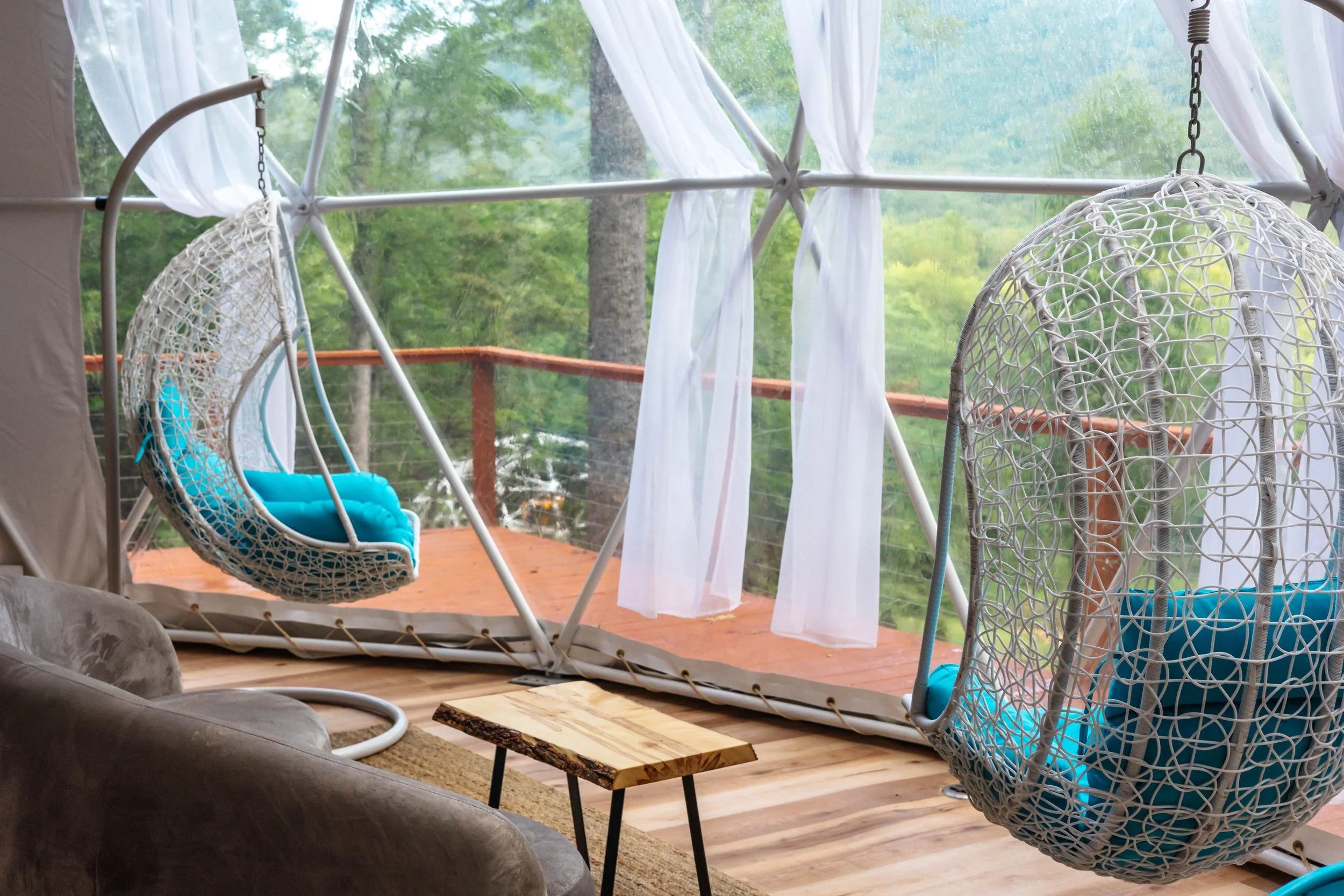 Indoor porch with two woven hanging chairs with blue cushions, a small wooden table, and a couch, with large windows and white curtains revealing a lush green outdoor view.