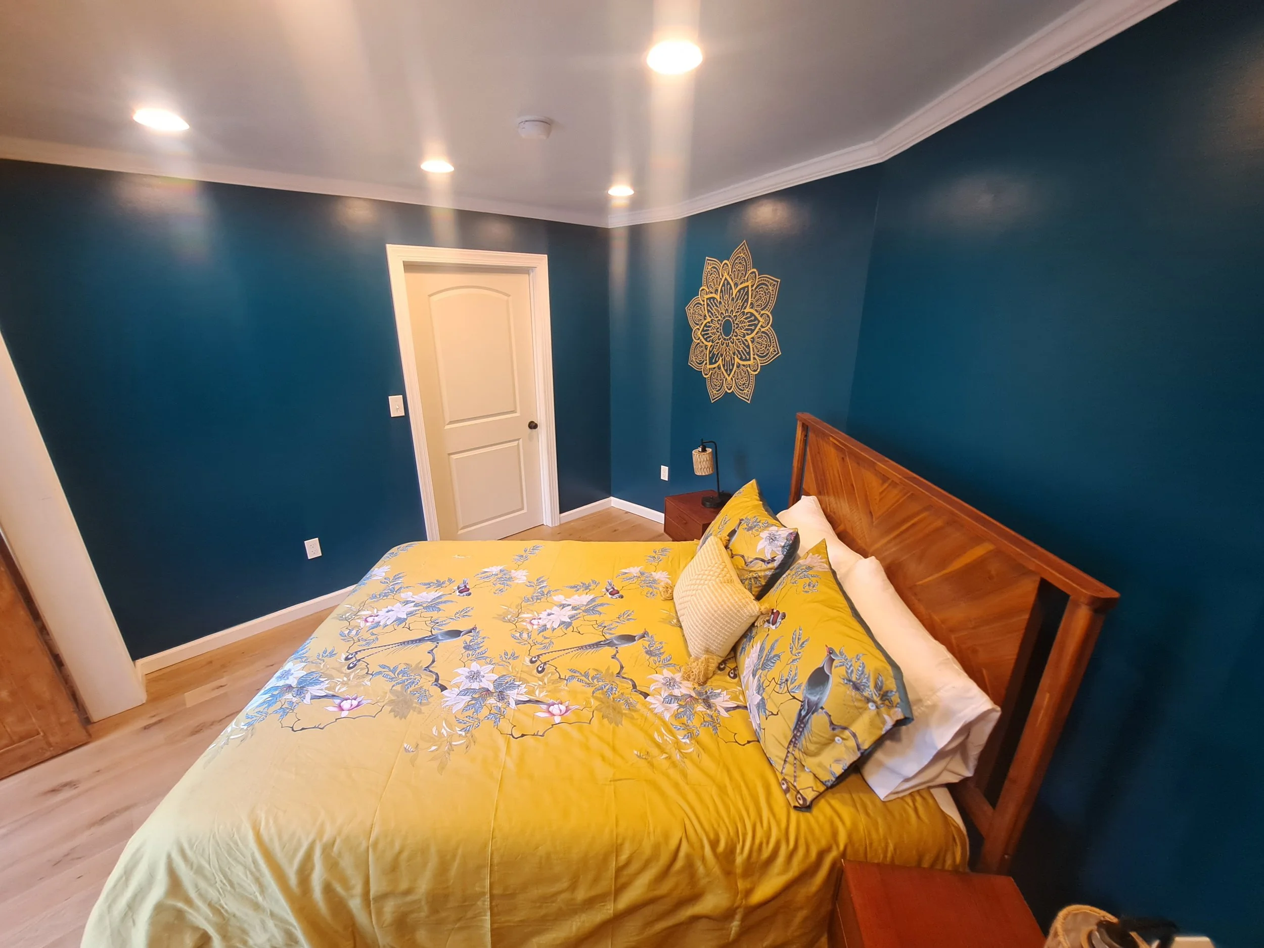 A bedroom with blue walls, a wooden bed with yellow and floral bedding, a small nightstand, and a wall decoration. Recessed lighting on the ceiling.