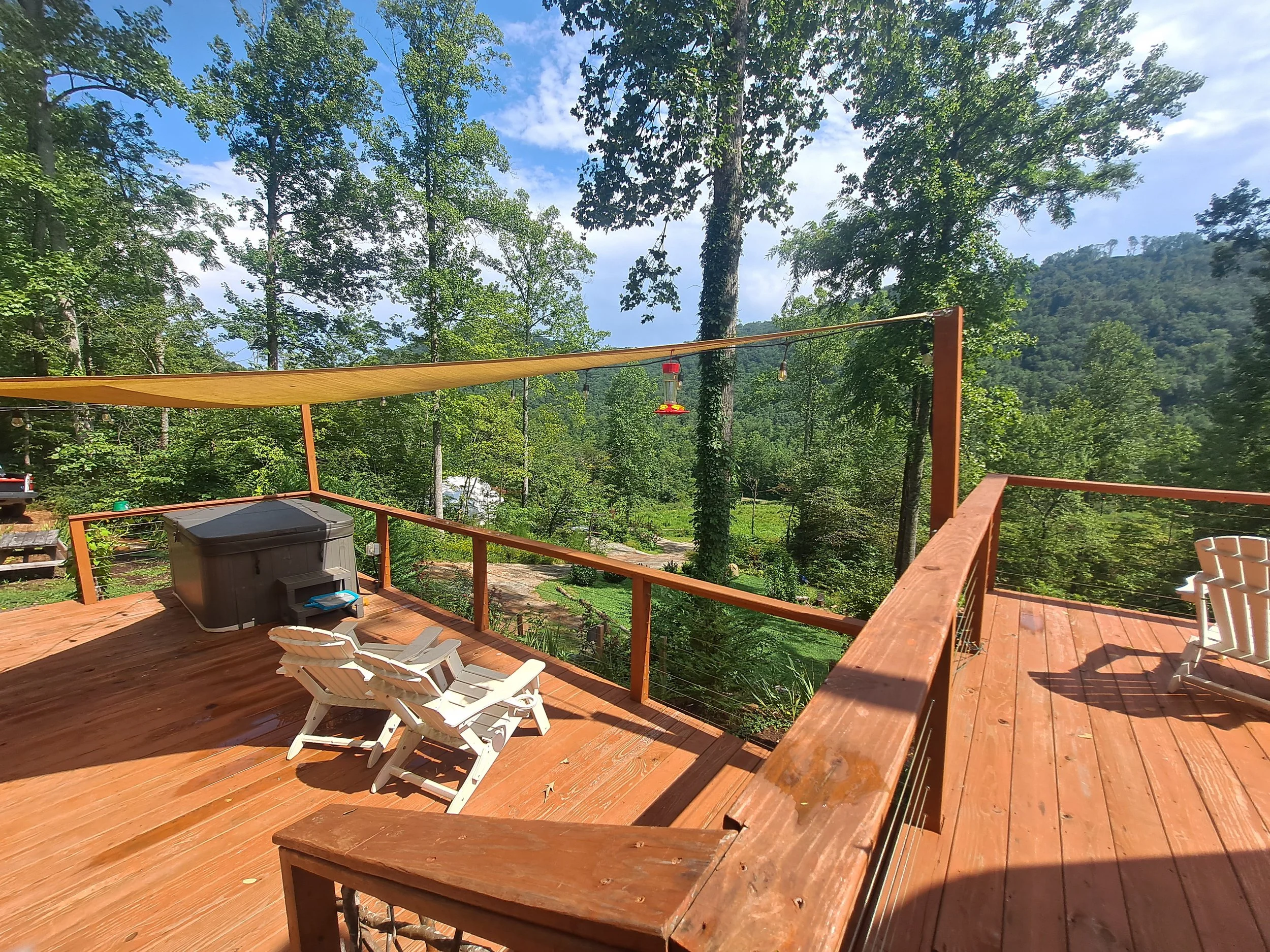 Wooden deck with two white lounge chairs, a hot tub, and a view of trees and mountains in the background under a partly cloudy sky.