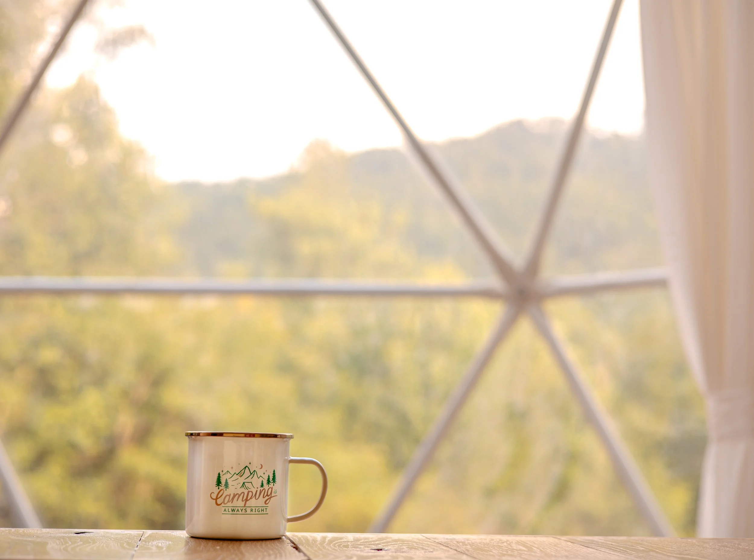 Camping mug with mountain and tree design and the words 'Camping Always Right' placed on a wooden surface, with a window showing an outdoor view of trees and mountains in the background.