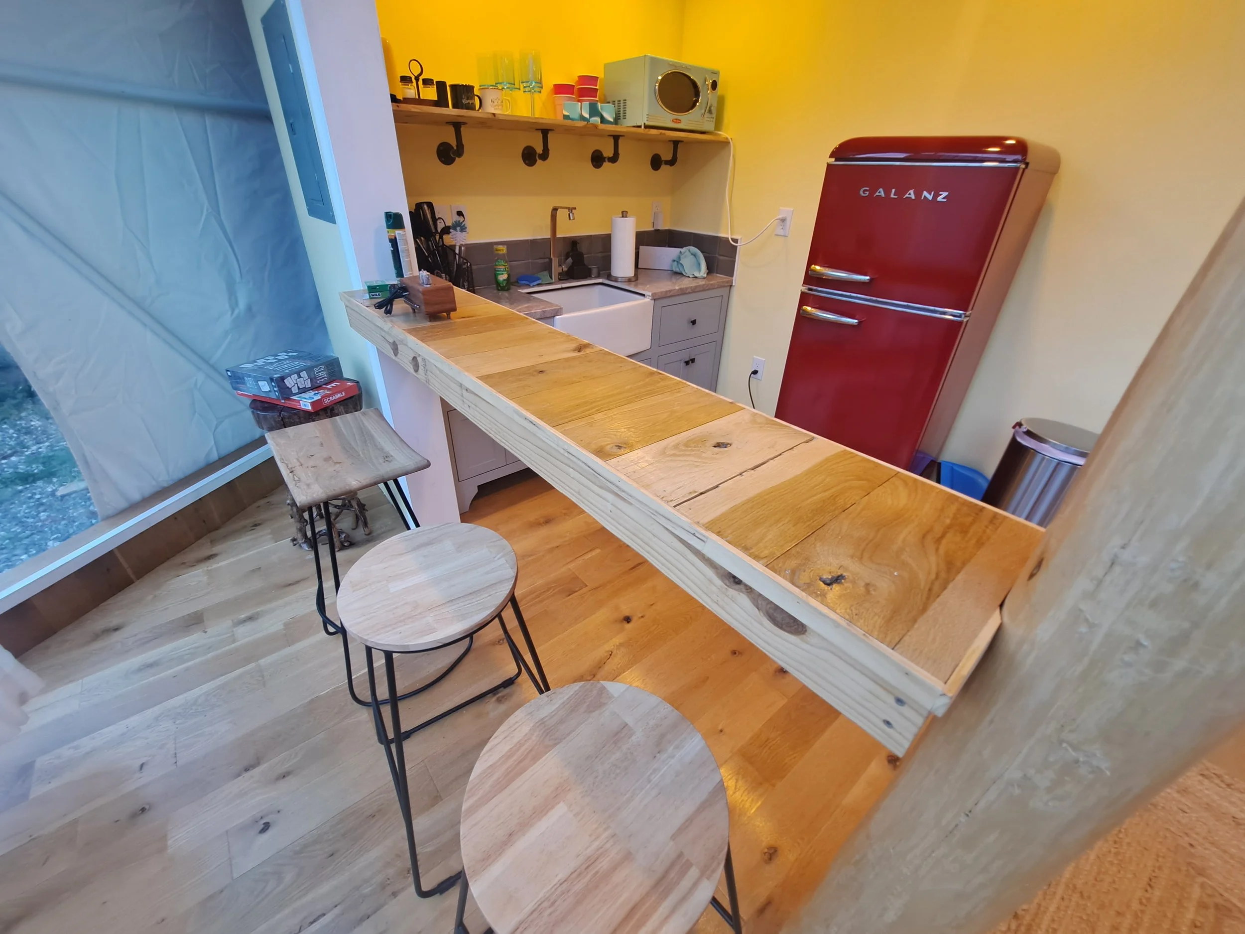 Interior of a small kitchen with a wooden bar counter and bar stools, yellow walls, a red vintage-style refrigerator, gray cabinetry, and open shelving with dishes and appliances.
