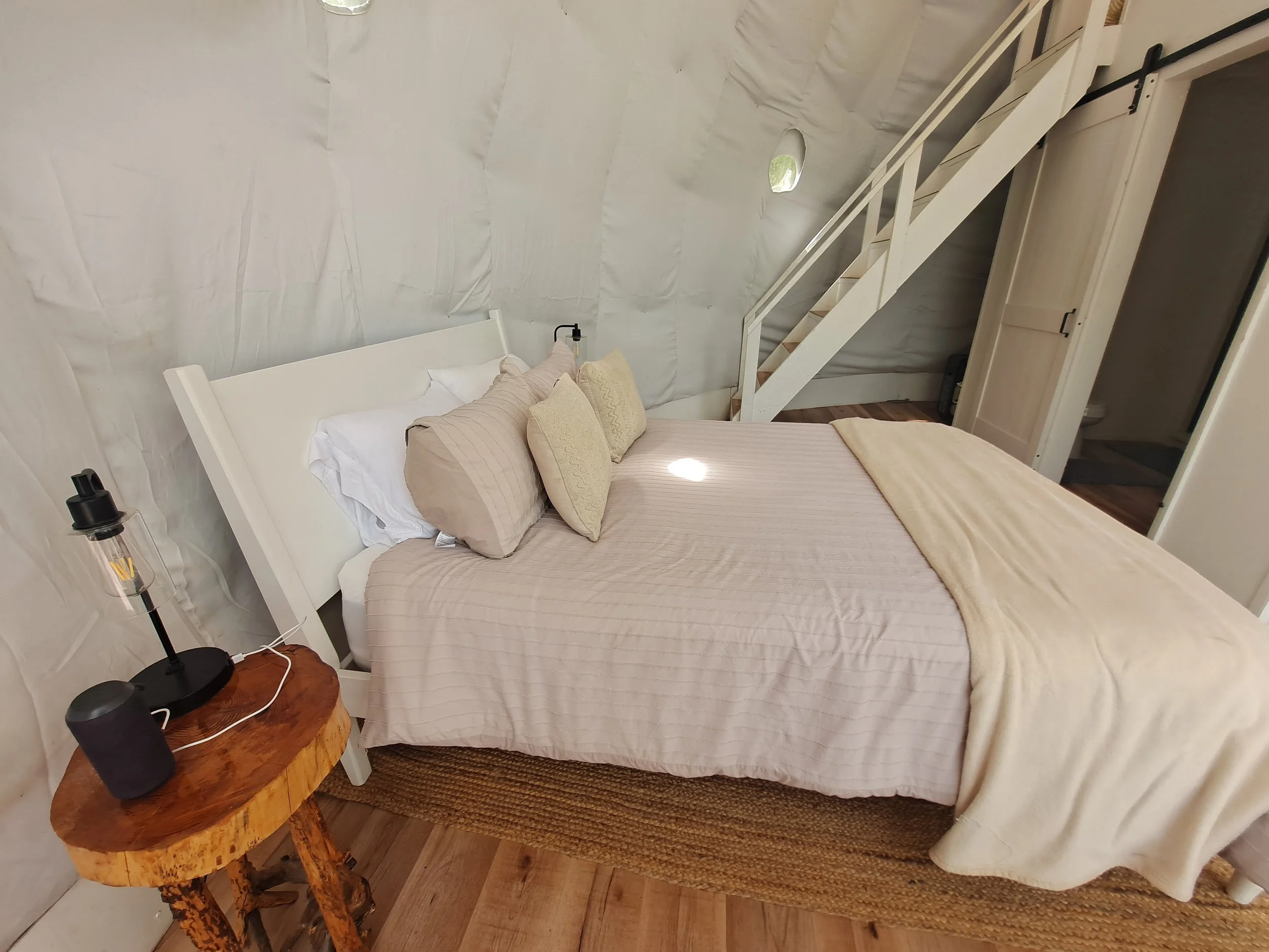 Bedroom with a white bed and beige bedding, pillows, and a blanket. Small round wooden side table with a black speaker and a lamp. Wooden staircase leading up, closet door, and small holes in the wall for ventilation.