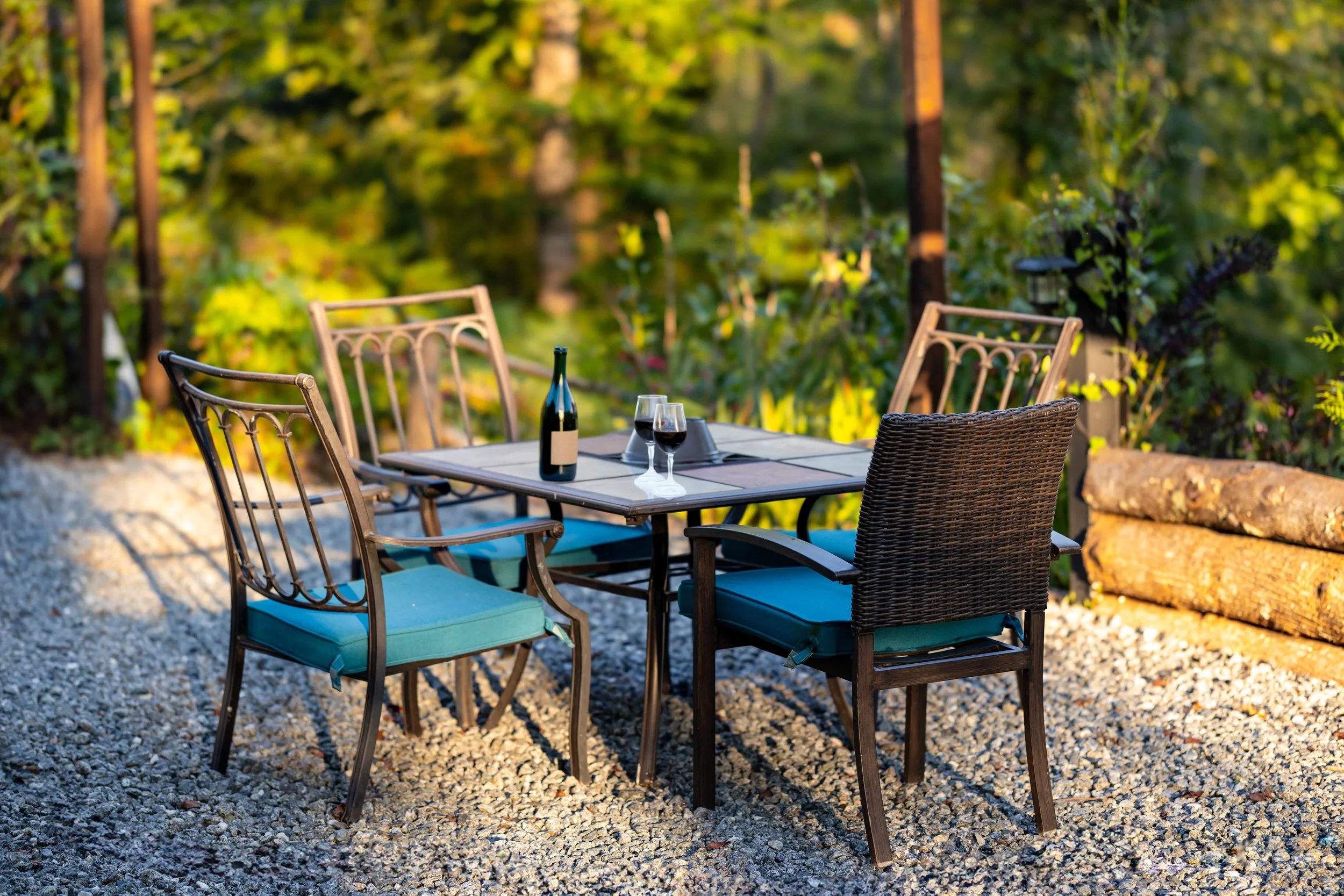 Outdoor dining table with four chairs, two wine glasses, a bottle of wine, and a small decorative item on it, set on a gravel surface with lush greenery in the background during daytime.