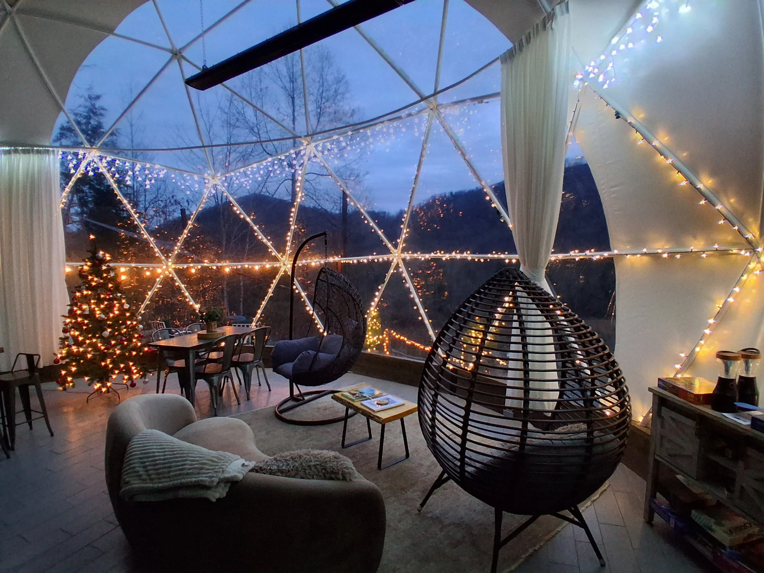 Cozy indoor space with string lights and a decorated Christmas tree, overlooking a mountain landscape at dusk.