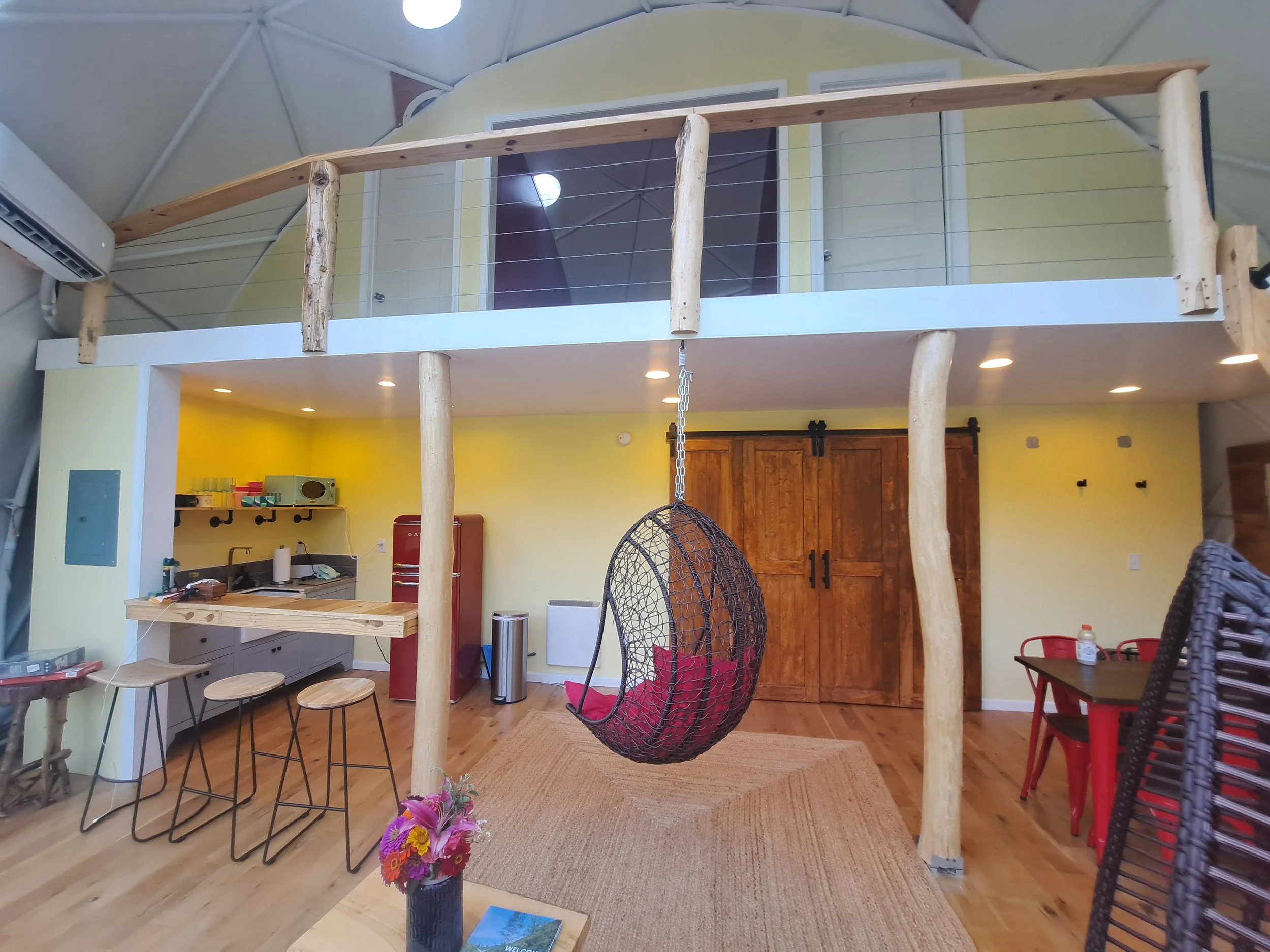Interior of a cozy, rustic-style room with a loft. The ground floor features a small kitchen counter with stools, a red refrigerator, and a wooden wardrobe. There is a hanging woven egg chair with red cushions in the center. To the right, a small tab
