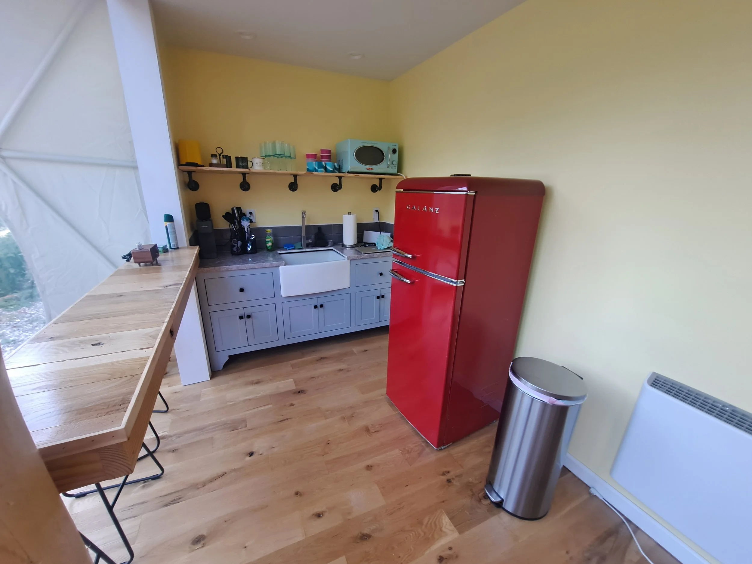 A small kitchen with yellow walls, a red refrigerator, a light blue cabinet with a white farm sink, and a wooden counter on the left side. There is a stainless steel trash can and a white heater on the right side, with shelves holding boxes, cups, an