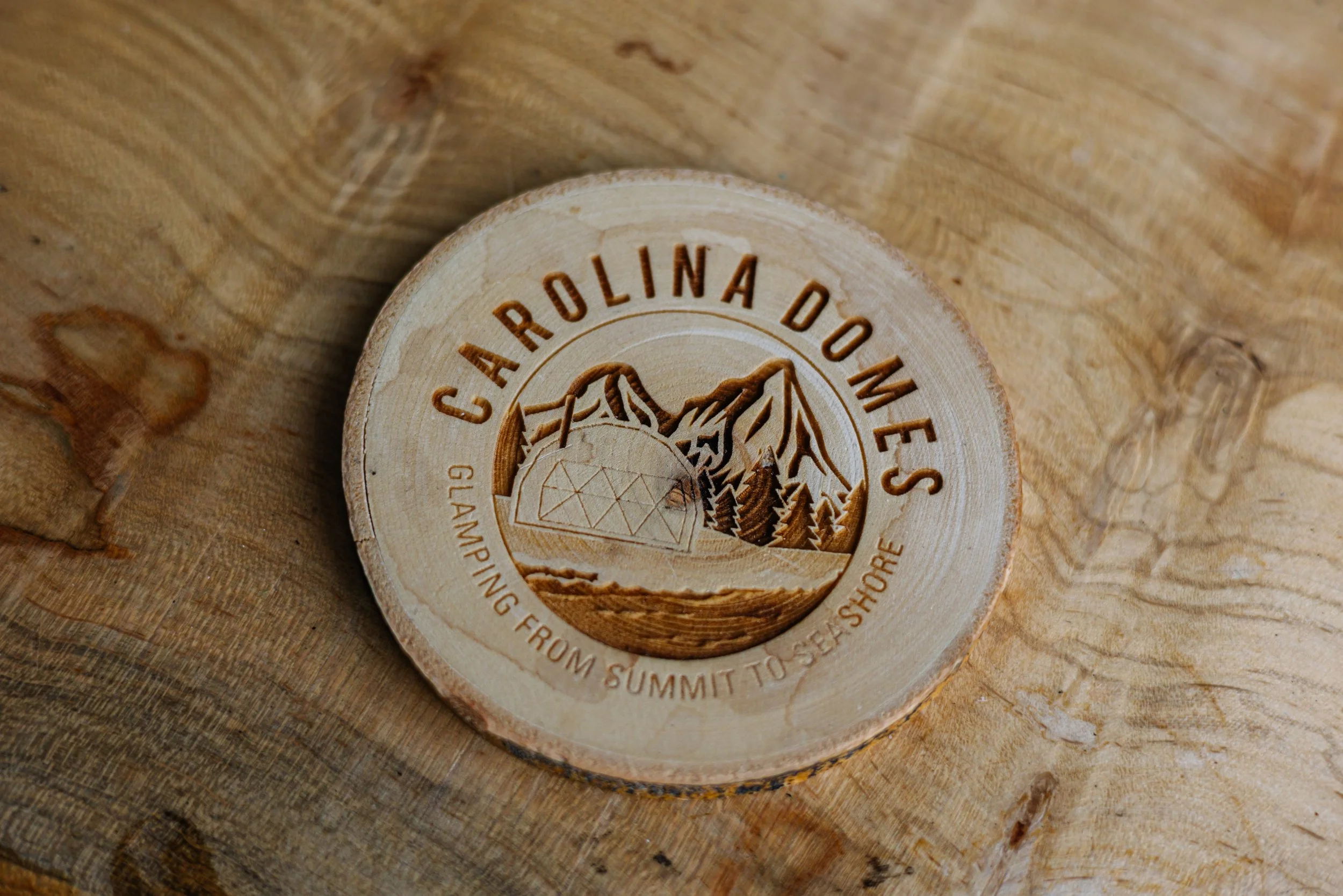 A round wooden emblem engraved with mountains, trees, a tent, and the words "Carolina Domes" and "Glamping from summit to sea shore," placed on a wooden surface.