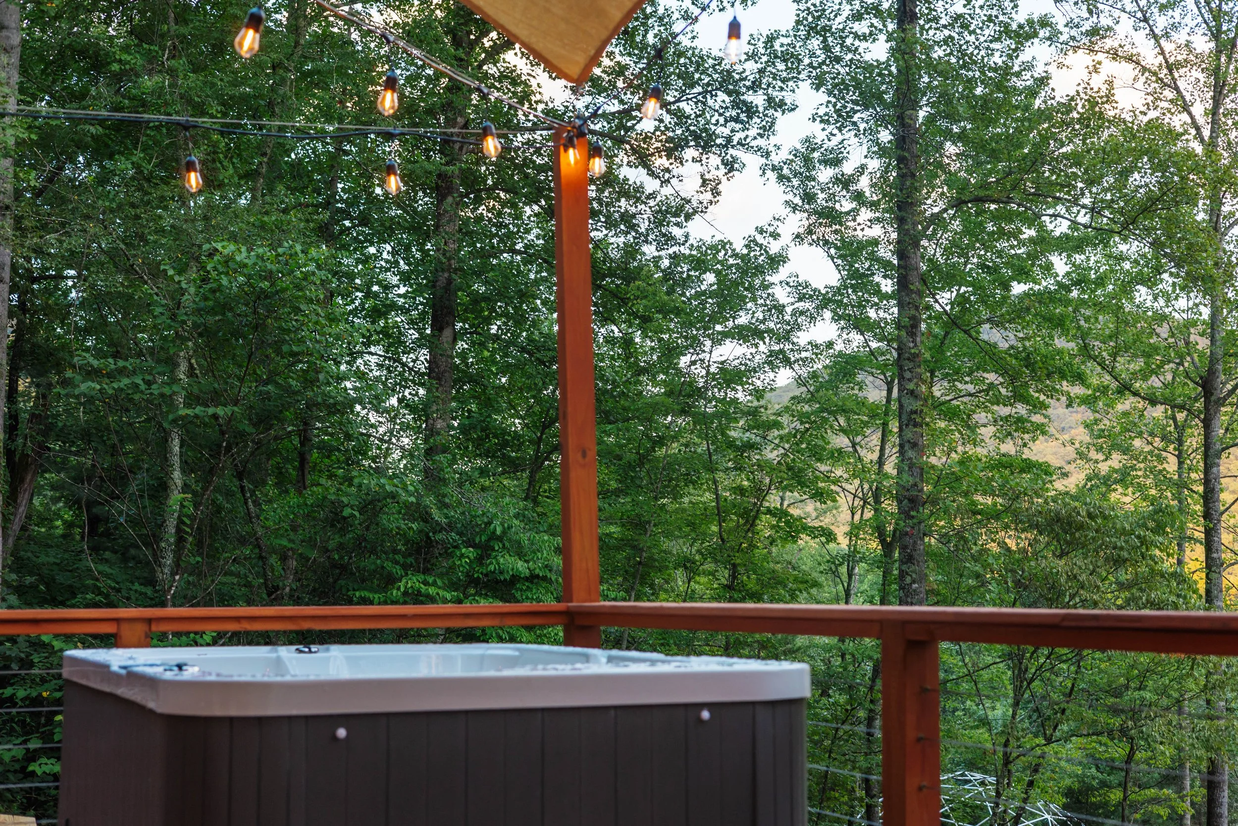Hot tub on a wooden deck with string lights overhead, surrounded by green trees.