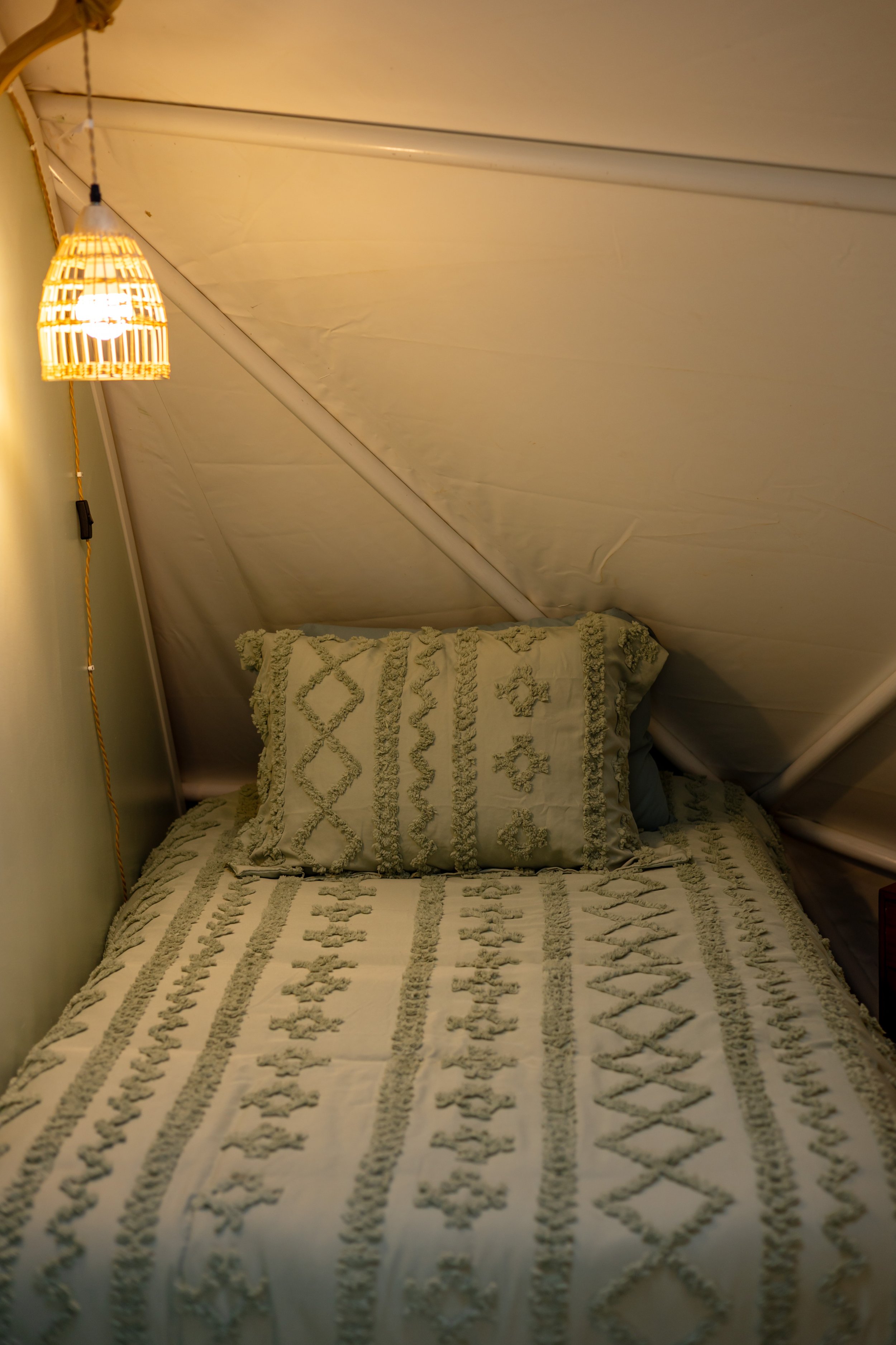A neatly made bed with an embroidered pillow and matching bedspread in a cozy, softly lit room with a sloped ceiling and a wicker lampshade hanging from the ceiling.