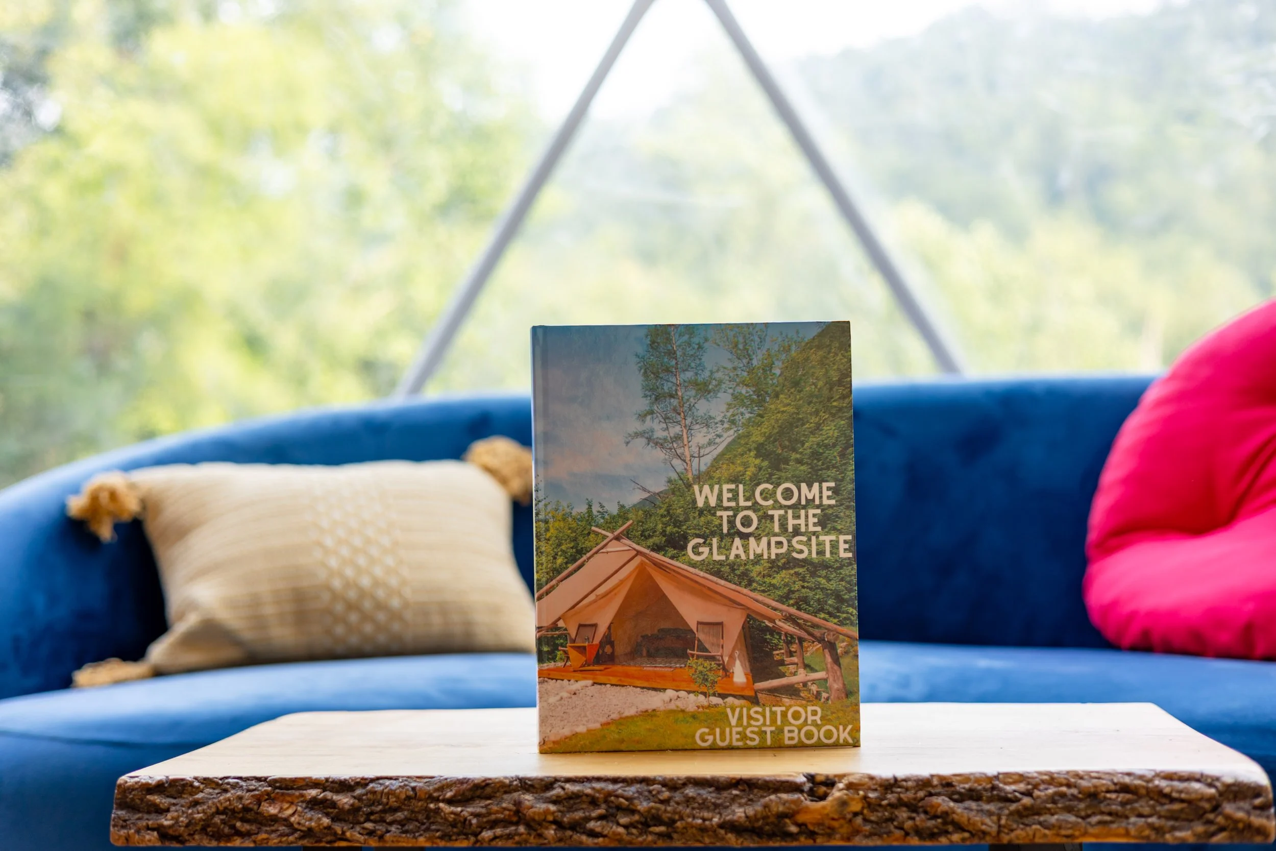 Visitor guest book on a wooden table at a glamping site, with a blue couch, pillow, and cushion in a cozy interior, and a blurred outdoor view of trees in the background.