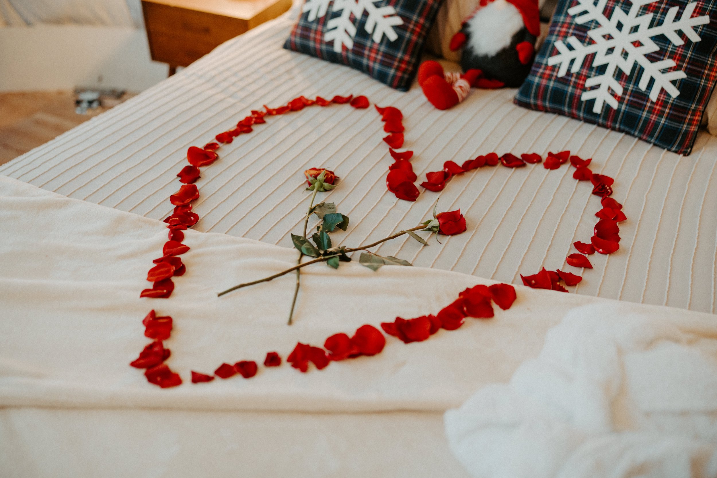 A bed decorated with red rose petals arranged in the shape of a heart. There is a single red rose in the center of the heart. The headboard has pillows with snowflake designs and a plush penguin toy.
