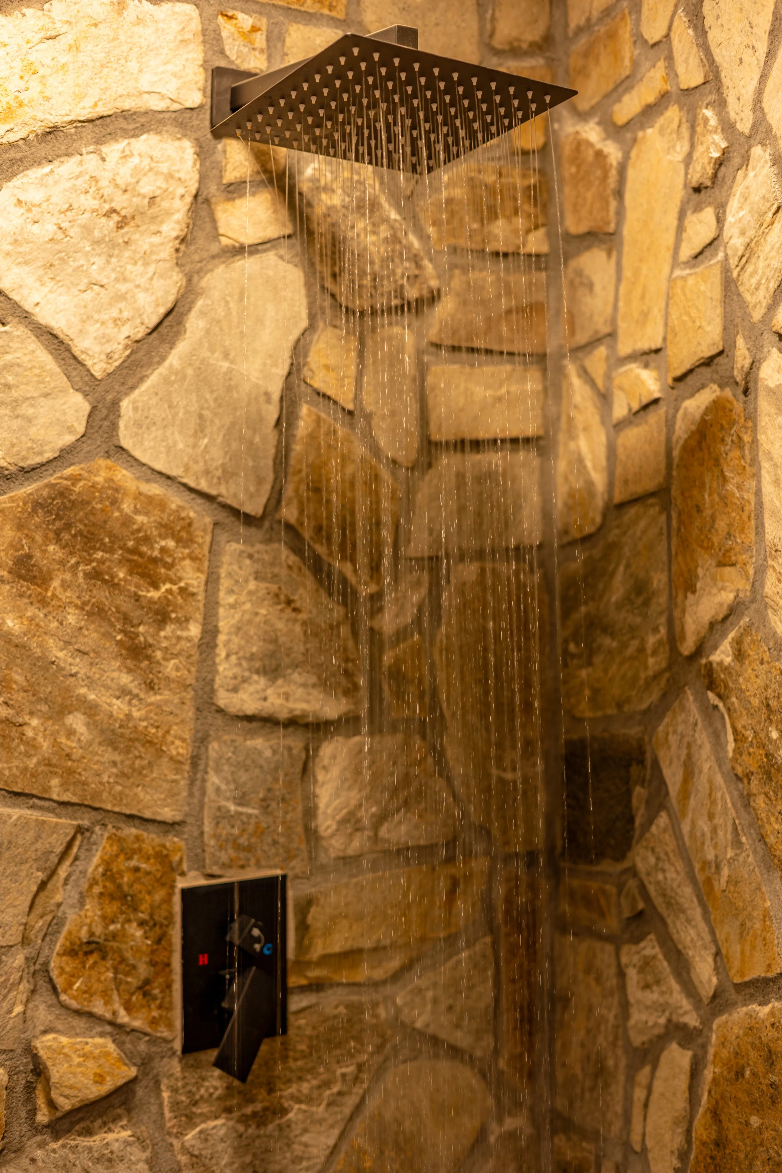 A modern shower with a square rain showerhead, water flowing, and a stone wall background.