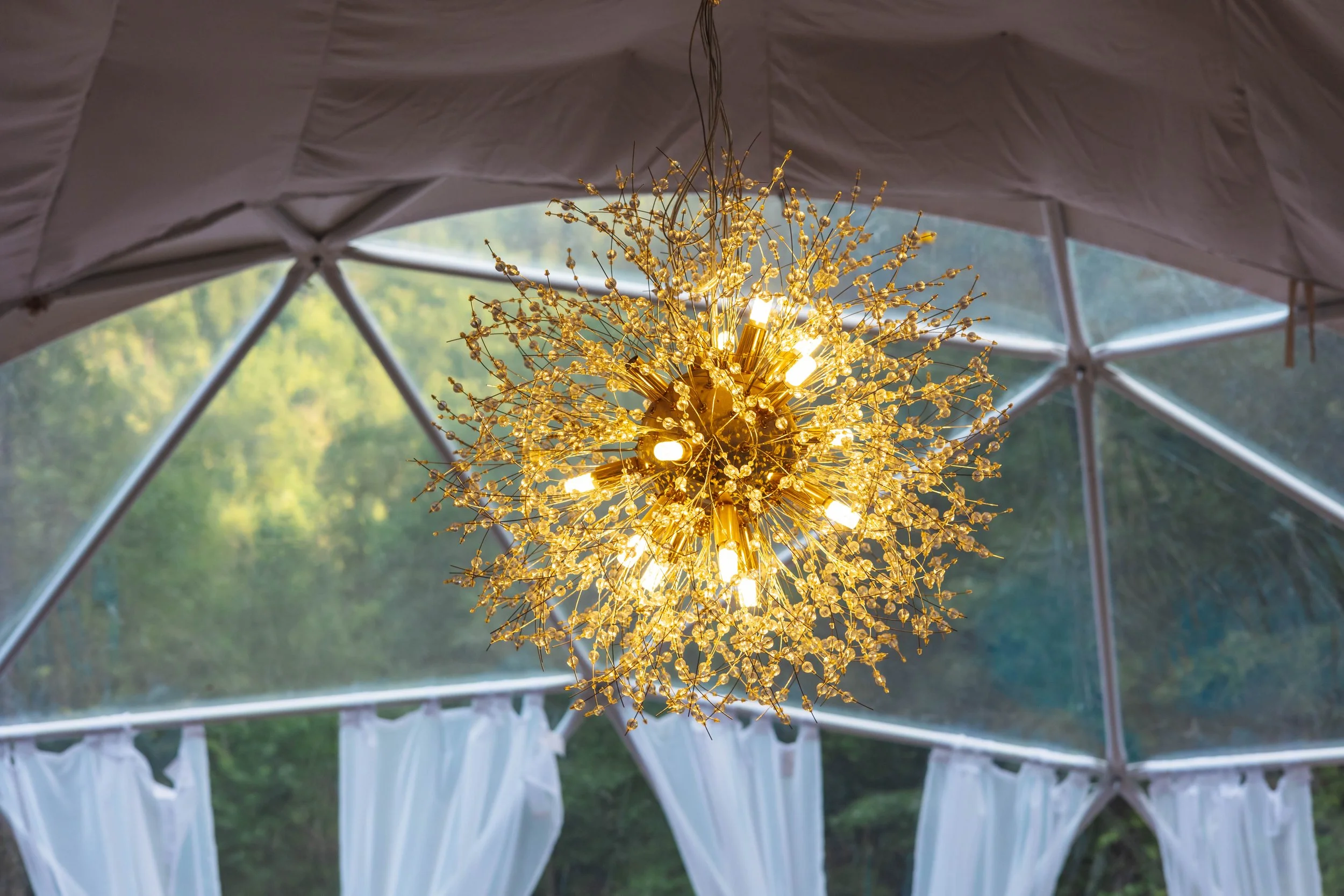 A modern chandelier with multiple small lights and gold decorative branches, hanging inside a tent with open sides and white curtains, overlooking a green outdoor landscape.